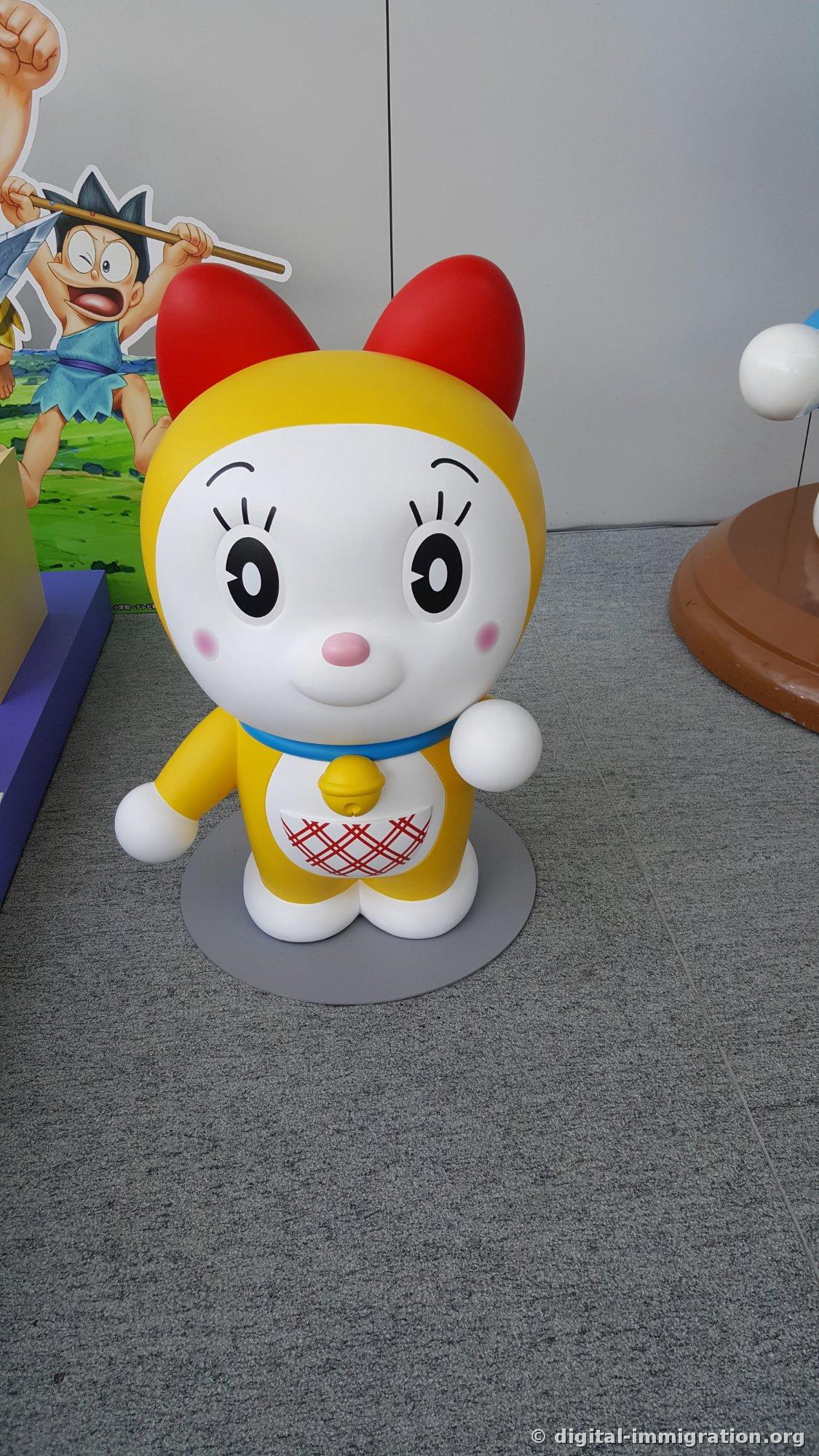 Figurine jaune de Dorami se dressant sur un tapis gris avec des rubans rouges et une grelot jaune, et un panneau publicitaire d'un film d'animation visible à gauche