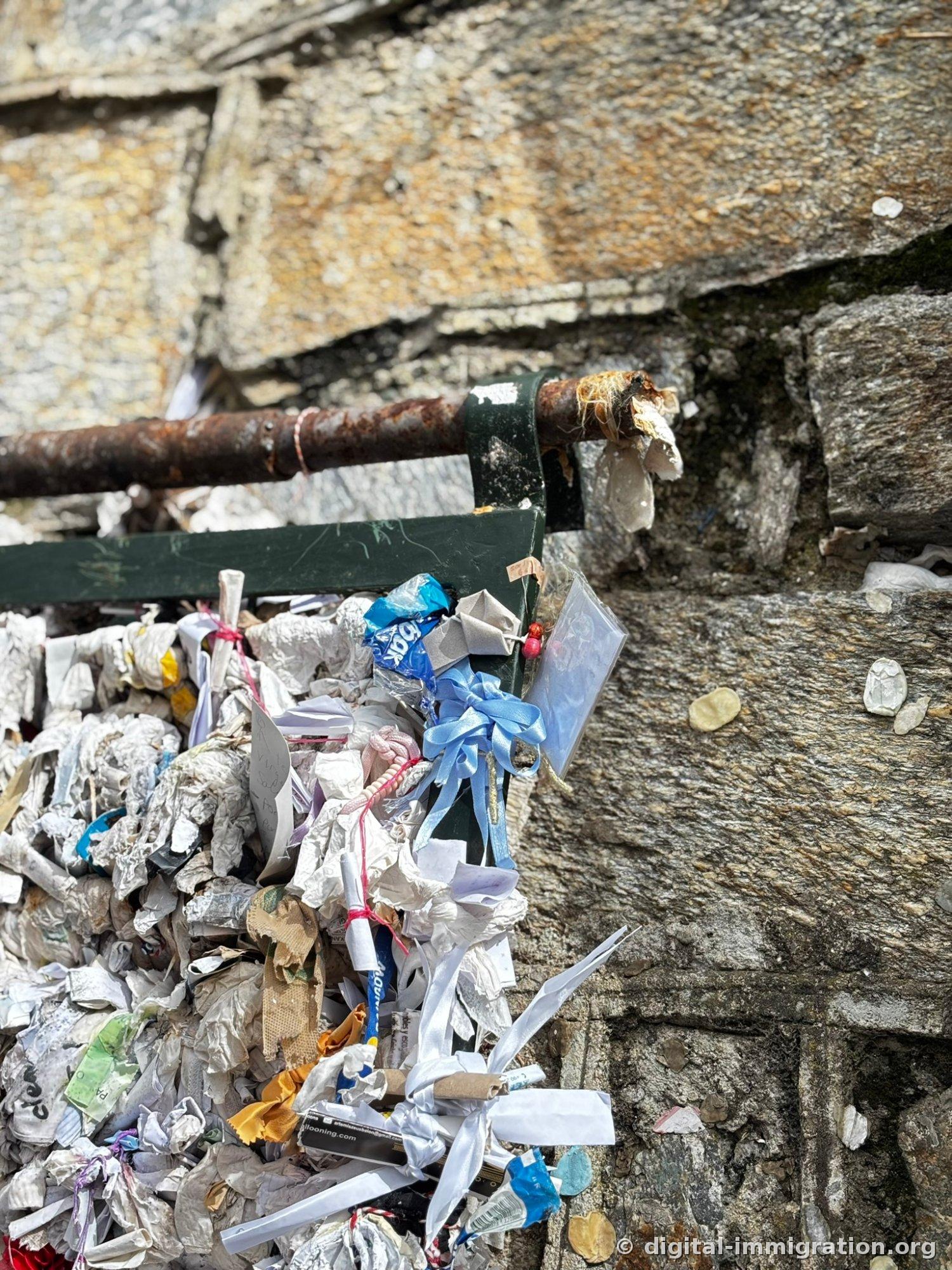 The end of the wish wall with a rusted iron pipe and green metal frame densely covered in paper knots, with a new light blue ribbon and a photo in a zipper bag hanging nearby