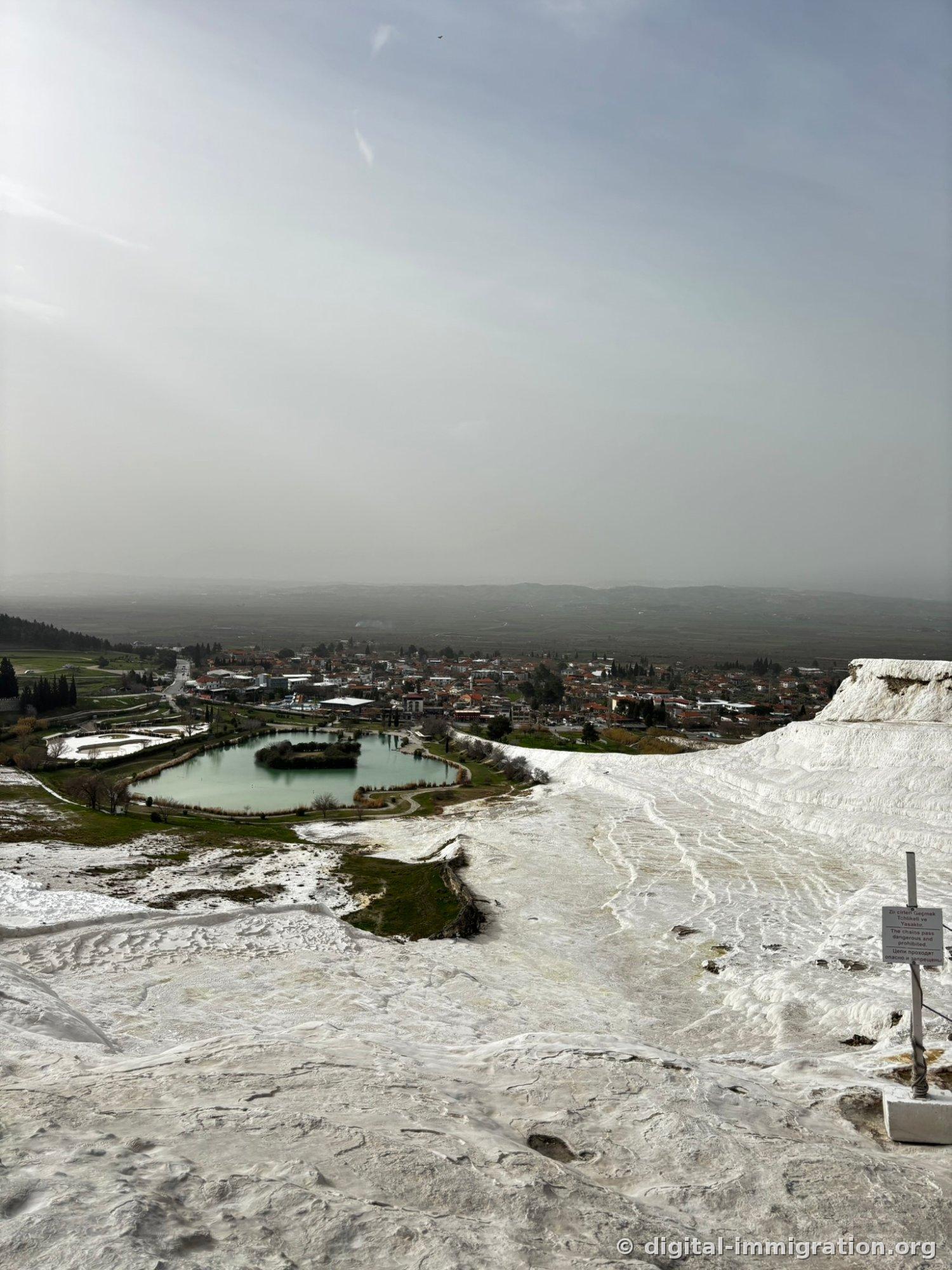 A view of the village of Pamukkale and green lake from the travertines, with a three-language warning sign on the right prohibiting entry past the chains