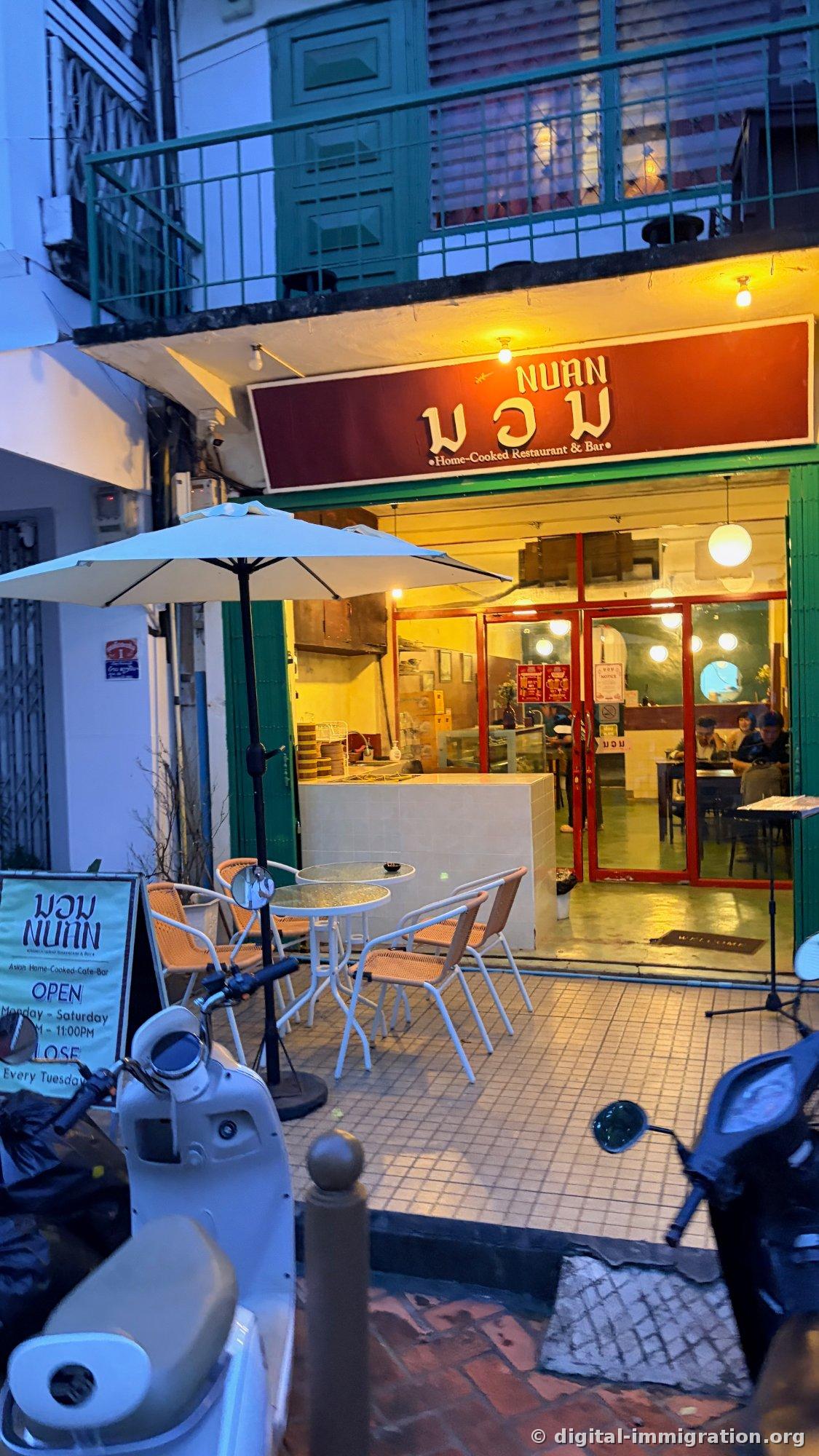 夕暮れ時に明かりがついたNUANレストランの外観。赤い看板にラオス語のນວນと『Home-Cooked Restaurant & Bar』の文字、前にはパラソルと屋外テーブル、スクーターが駐車されている