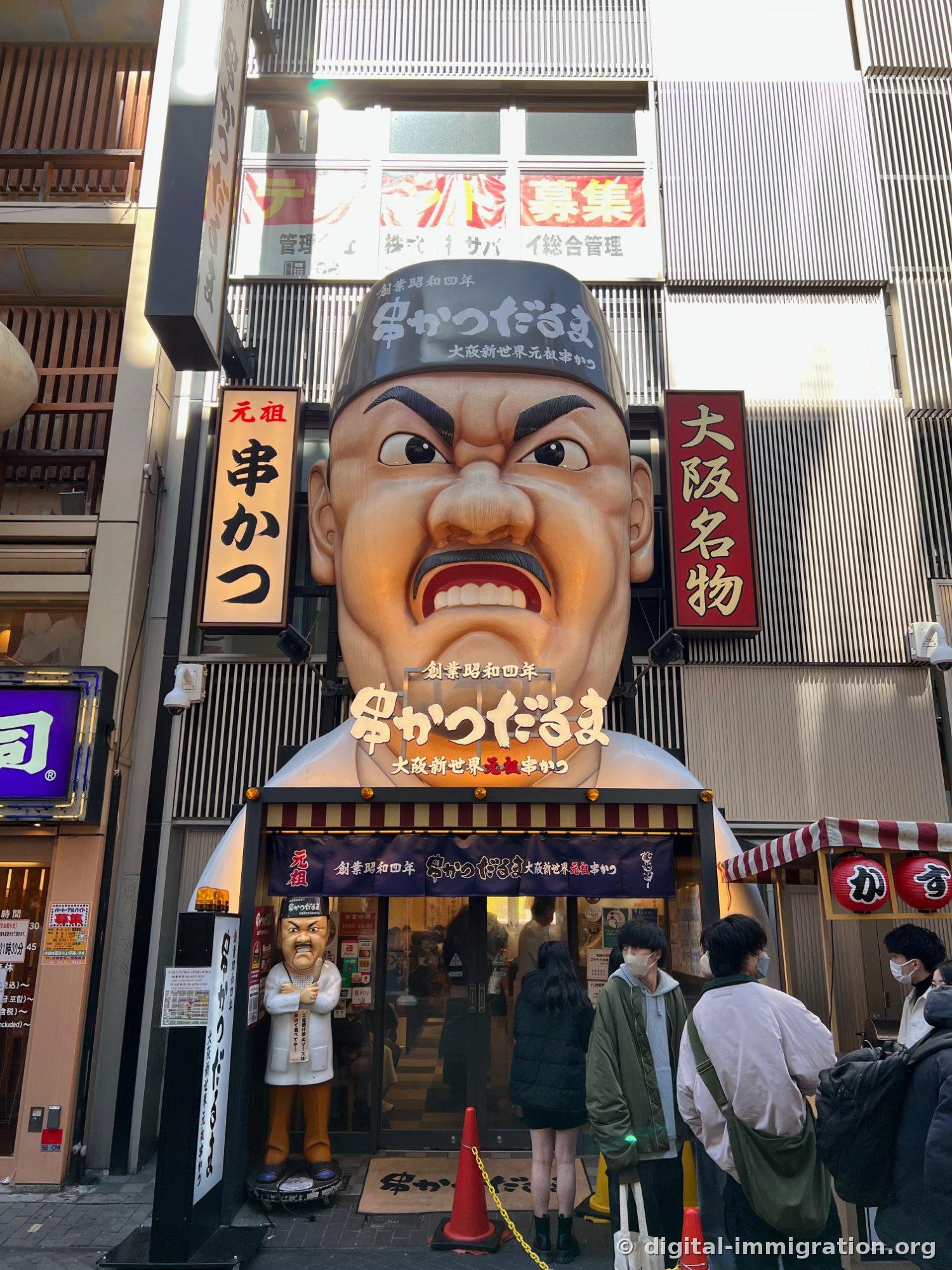 Énorme statue du visage en colère du vieil homme à l'entrée de la succursale principale du kushikatsu Daruma, avec une file d'attente de clients