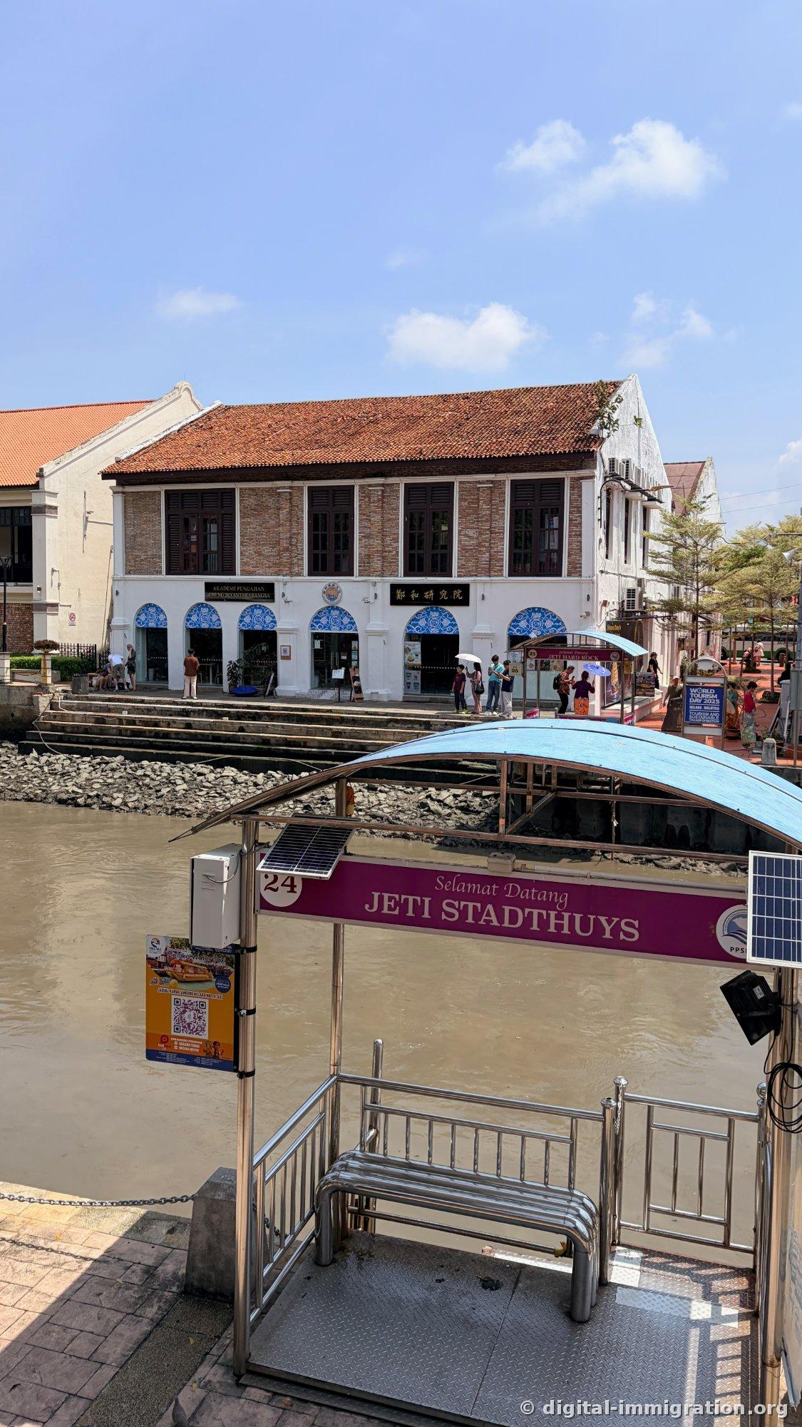 The river cruise dock with a blue roof and deep pink JETI STADTHUYS sign. The Cheng Ho Research Academy brick building is visible across the river with people holding white parasols