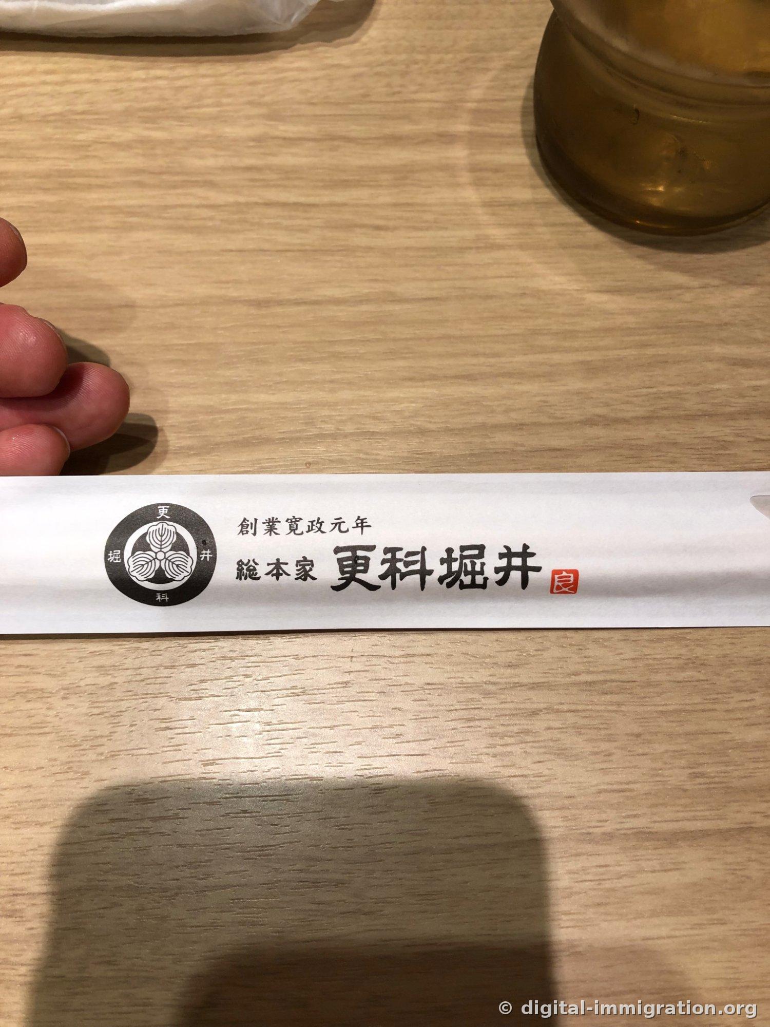 A white chopstick wrapper with 'Founded in Kansai Year One, Sarashina Horii Head Shop' and a three-leaf logo printed on a wooden table