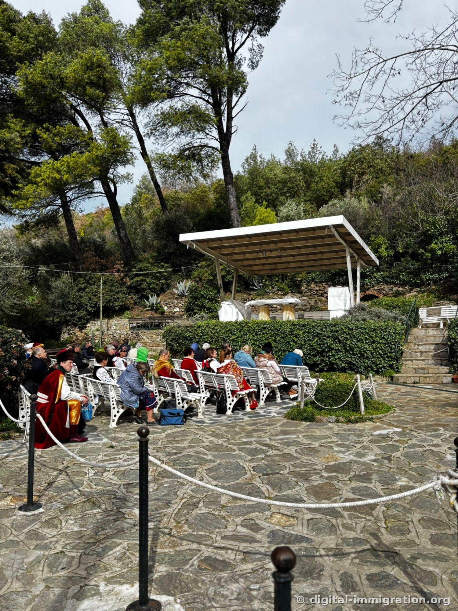 People in crimson velvet traditional dress trimmed with fur and feathered hats stand or sit on benches in a stone plaza, with a red SIGARA ICILMEZ (NO SMOKING) sign on the nearby railing