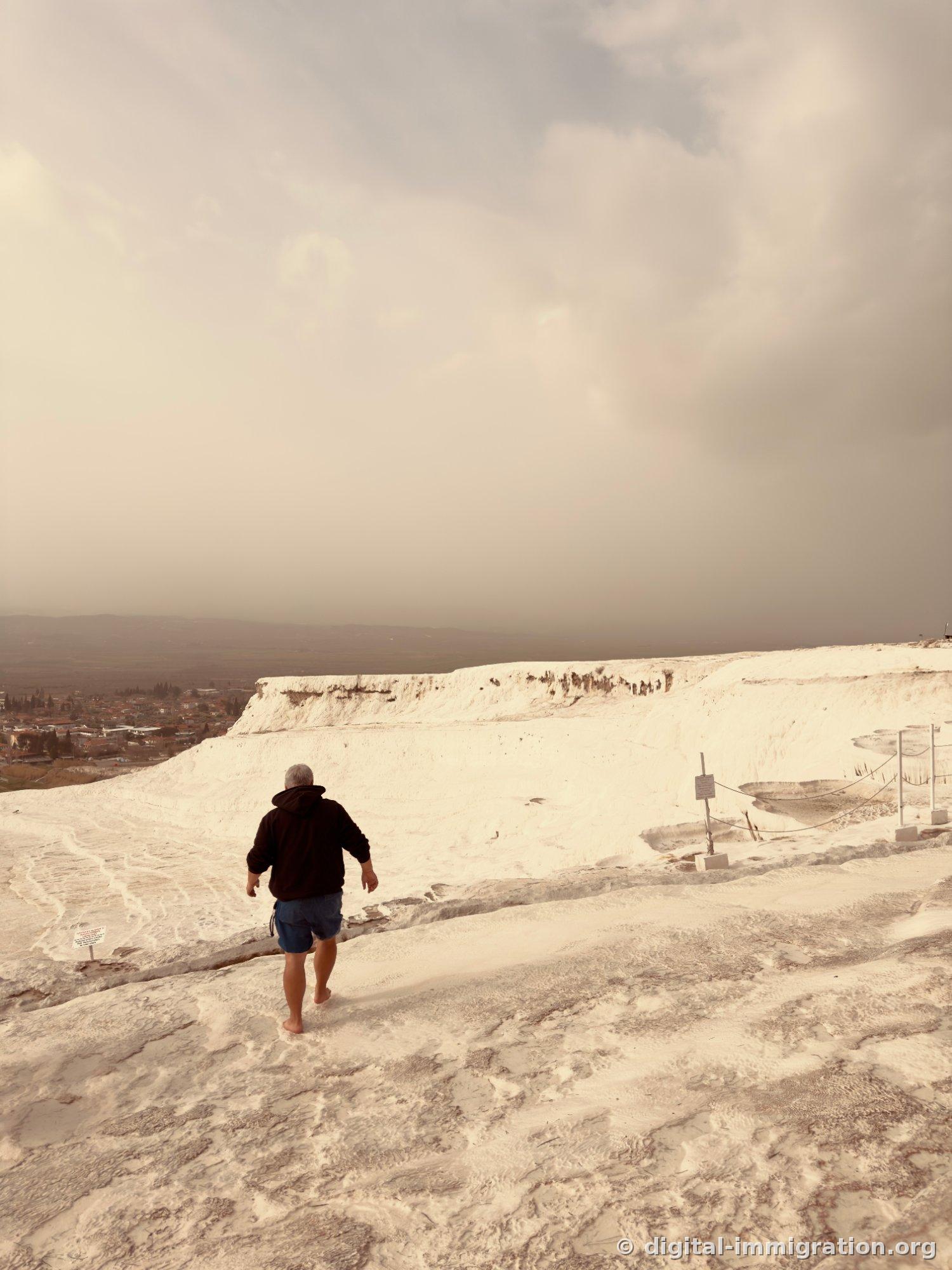 The back view of a man in shorts walking barefoot down a dry white limestone hill, with the village and overcast sky visible in the distance
