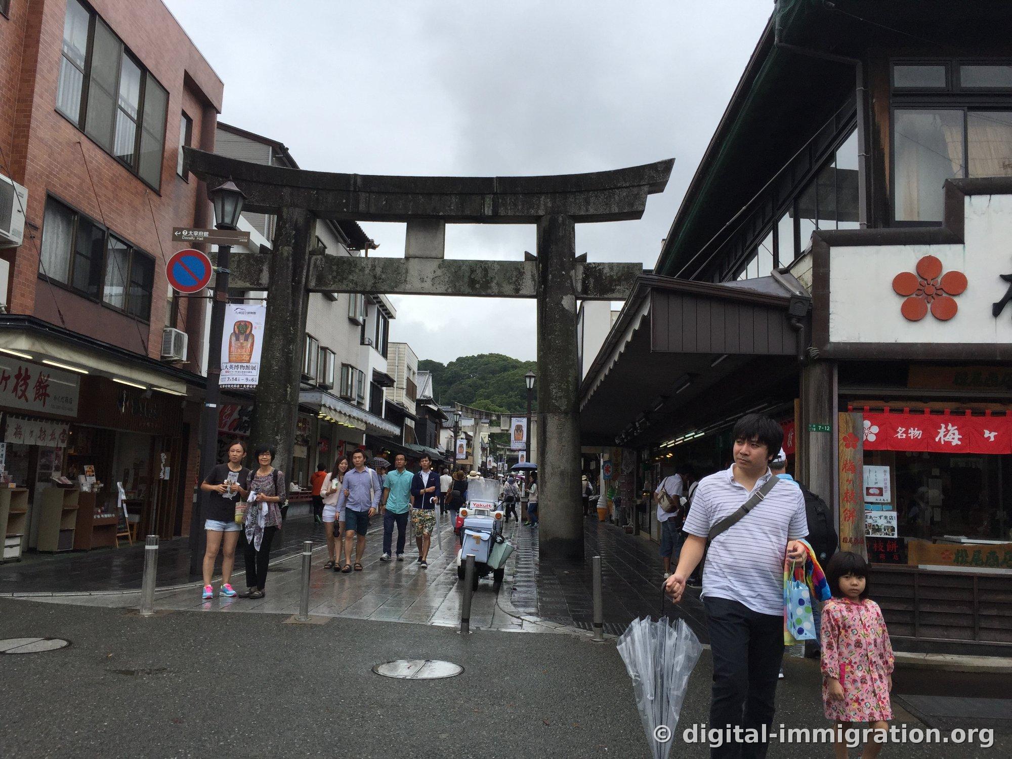 Stone torii standing in the middle of Dazaifu Sando shopping street with umegaemochi shops lined up on both sides, red plum blossom shop signs