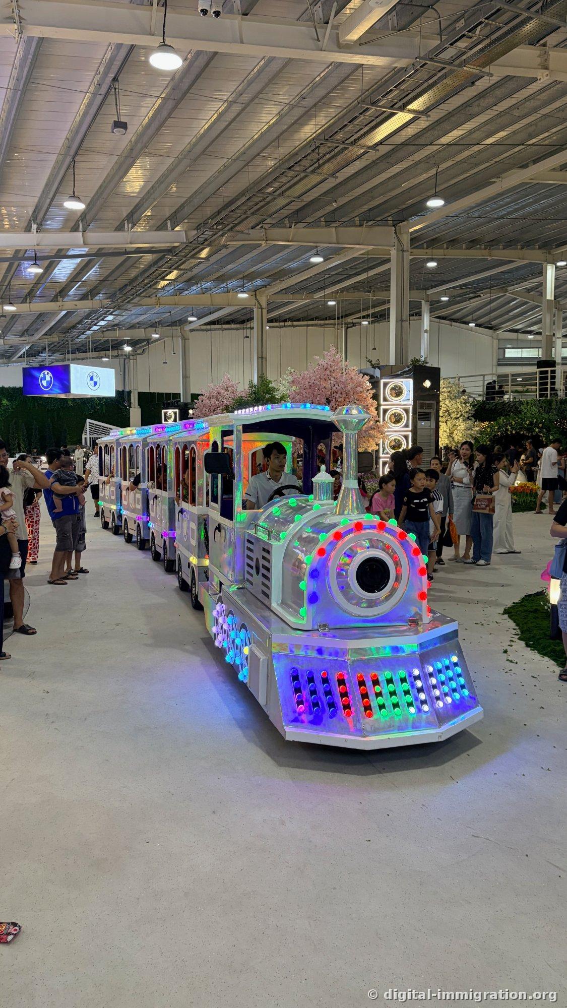 LED-lit miniature train with operator and children riding, families gathered watching, BMW sign in background