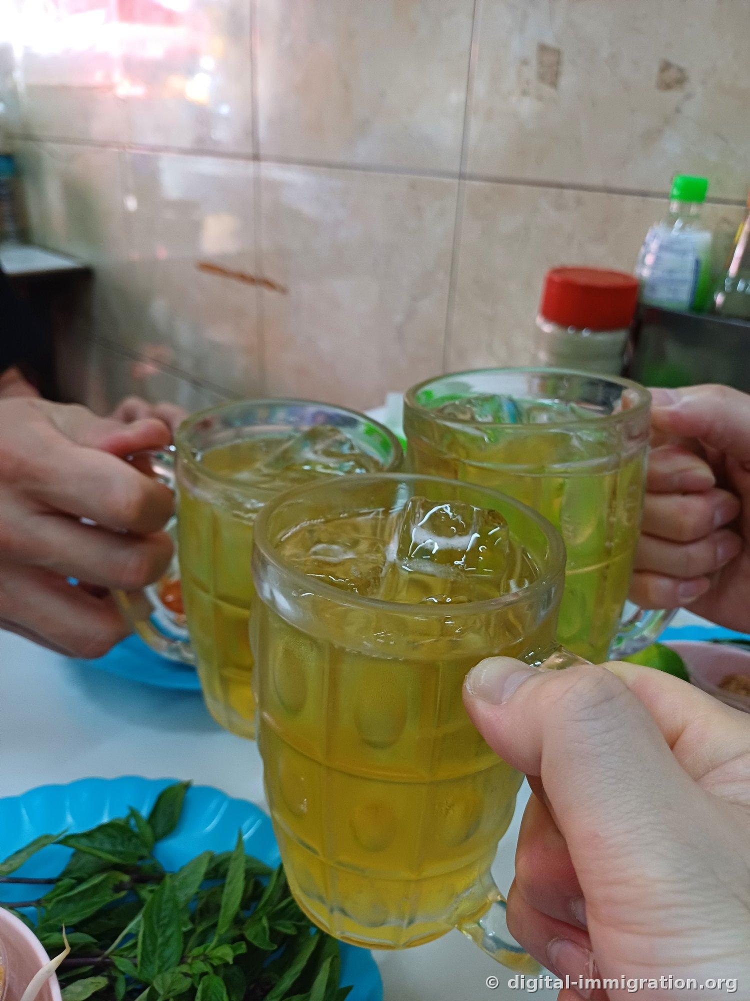 餐厅里三个人举着装冰的啤酒杯碰杯，前面蓝色盘子上放着罗勒叶，后面是瓷砖墙和调料瓶