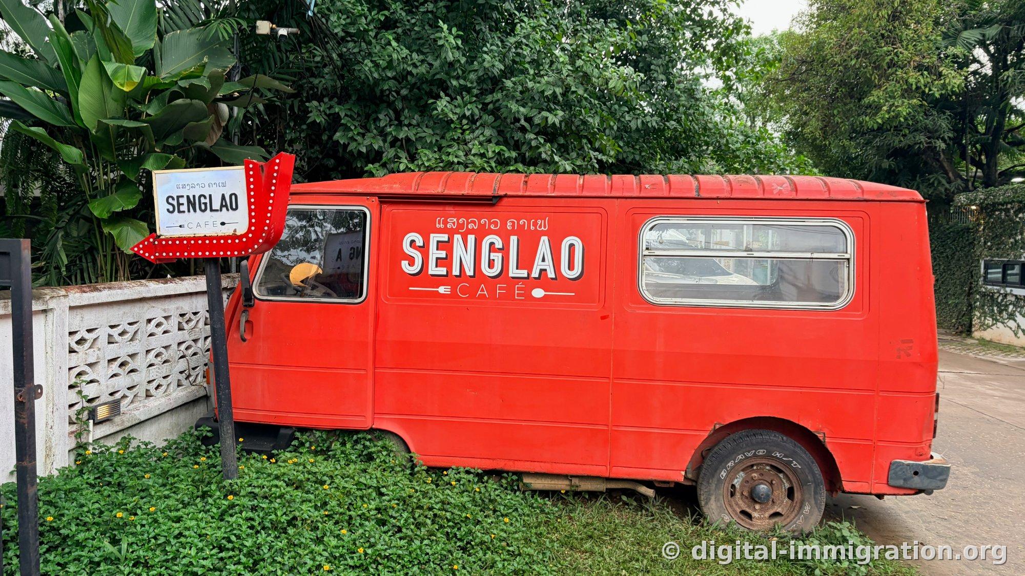 Un minibus rouge garé dans l'herbe au bord de la route avec le logo 'SENGLAO CAFÉ' en lettres blanches et une enseigne en laotien, une flèche rouge avec une ampoule à côté