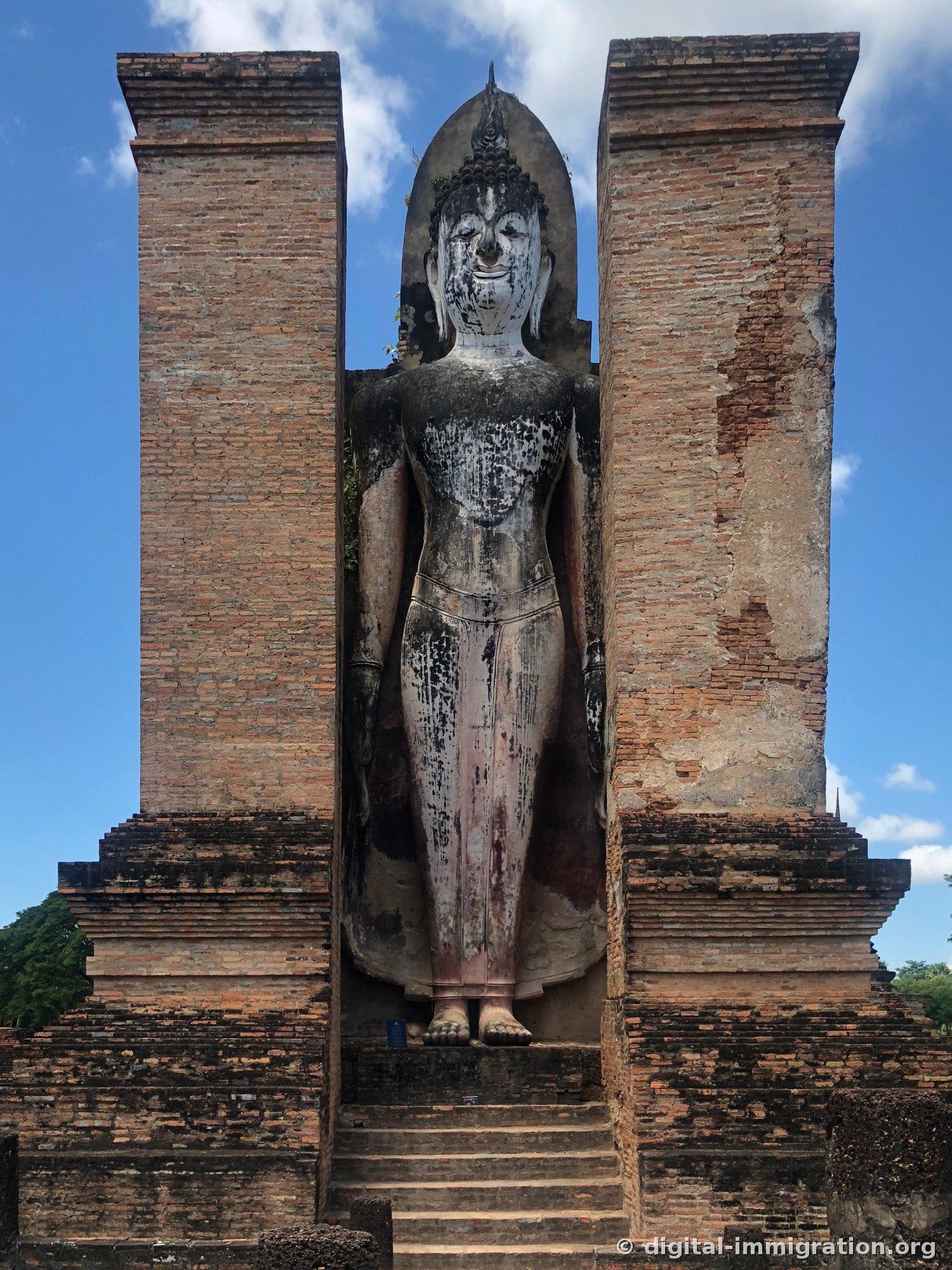 A giant standing Buddha between two brick columns, with black water stains flowing vertically down the upper body and stairs leading down below