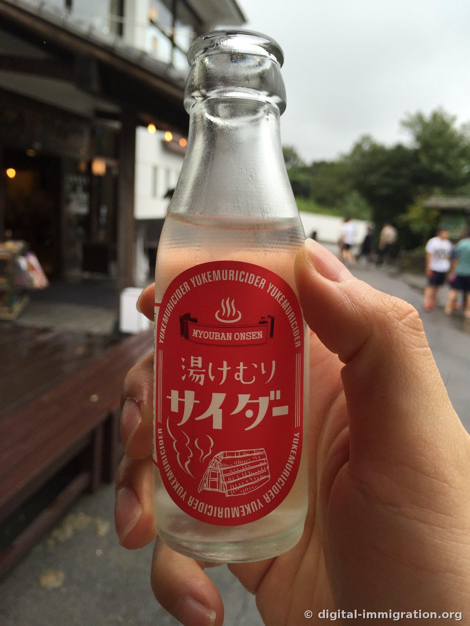 Une bouteille de verre de cidre avec un label rouge lisant 'MYOUBAN ONSEN 湯けむりサイダー' tenue à la main, avec un bâtiment de boutique en bois en arrière-plan