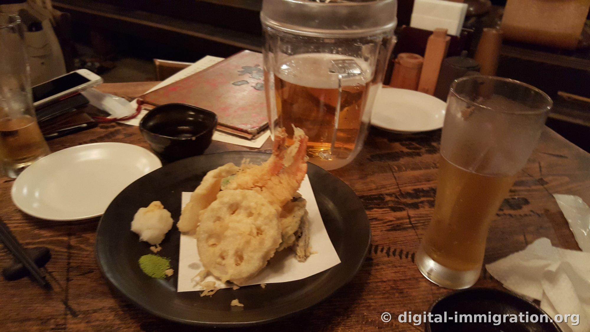 Shrimp, lotus root, and eggplant tempura on paper atop a black plate with grated daikon and matcha salt beside it, with a draft beer tankard, glass, and menu on a dark wooden table
