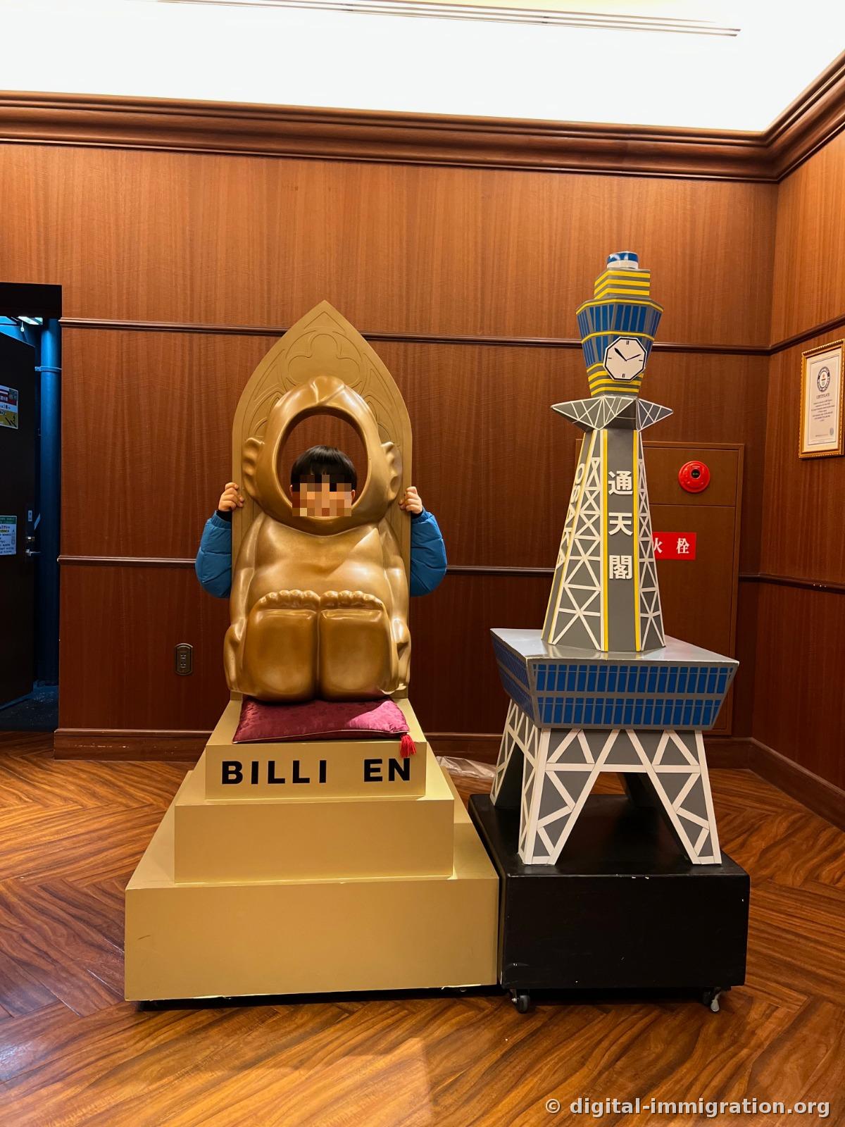 A child putting their face in a golden Billiken photo spot, with a Tsutenkaku model placed beside