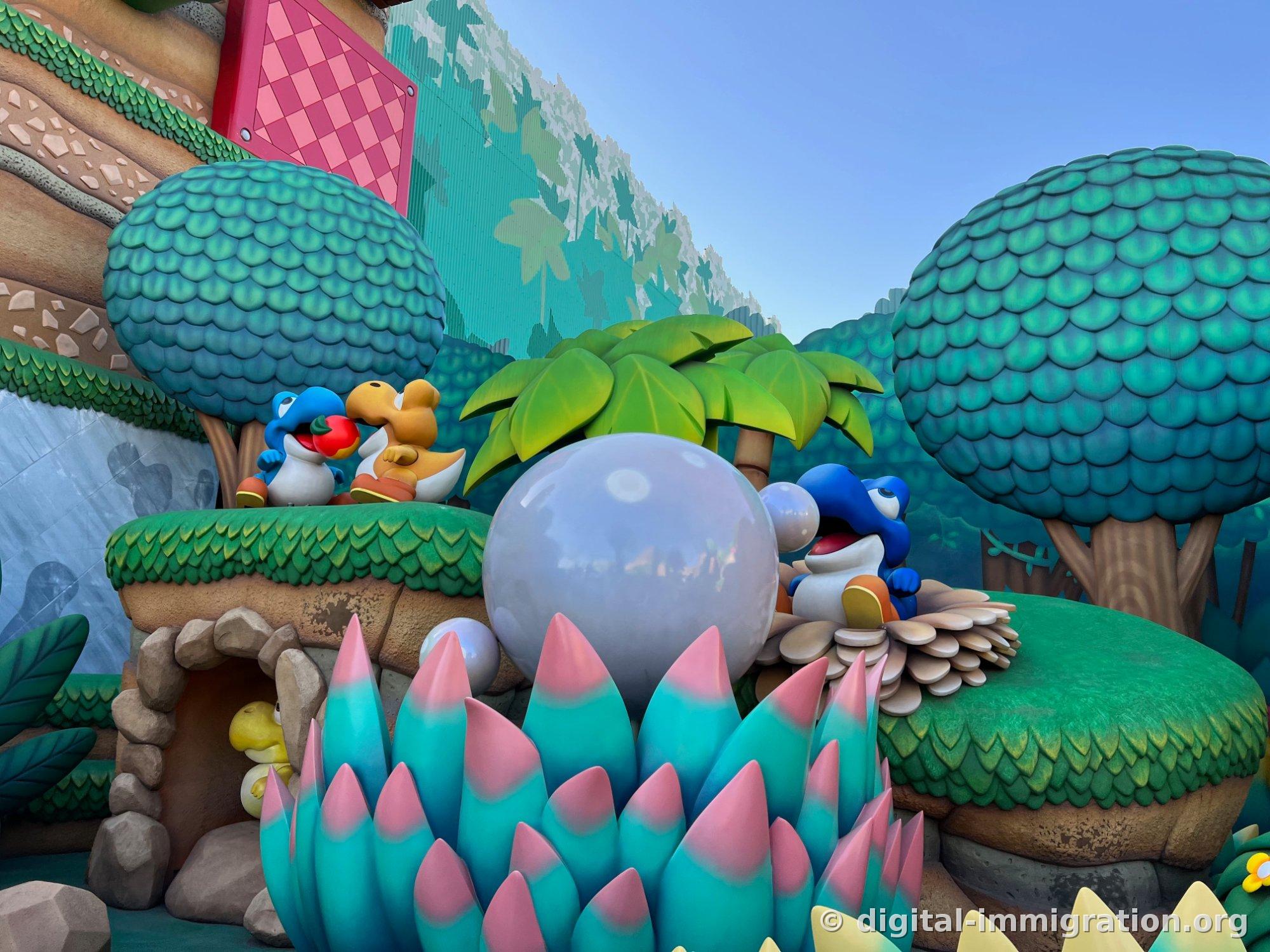 A blue Yoshi holds a red apple, with a yellow Yoshi and another blue Yoshi positioned on a green hill, with pink and light blue spiky plants and a large white sphere in front