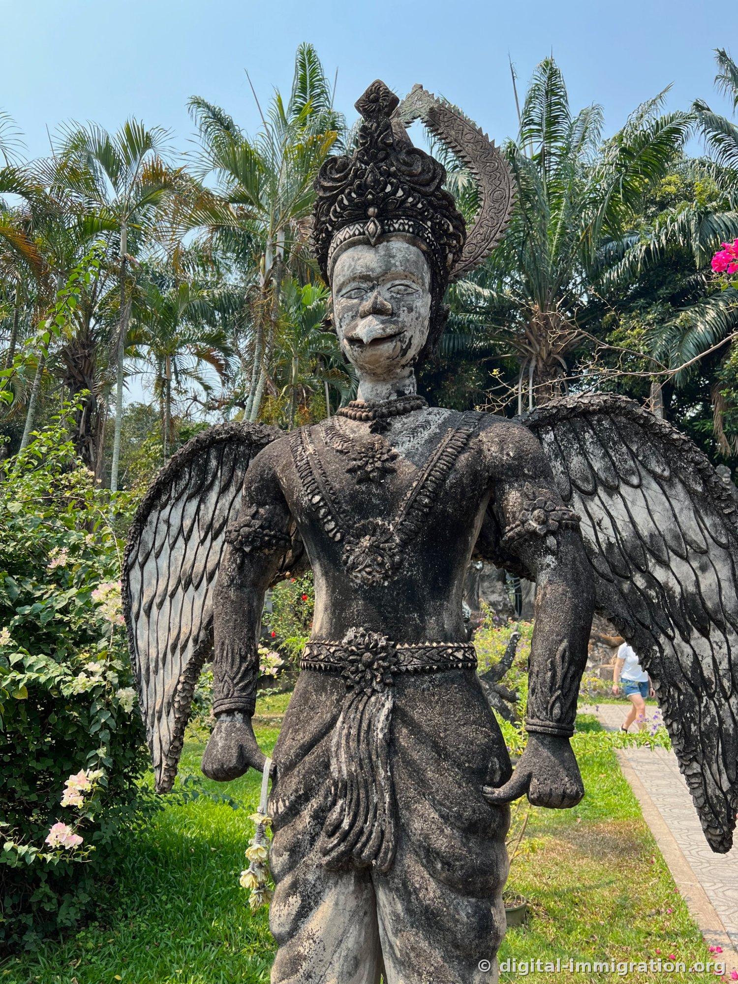 Une statue ailée qui s'est noircie avec le temps, portant une couronne élaborée, avec des palmiers en arrière-plan