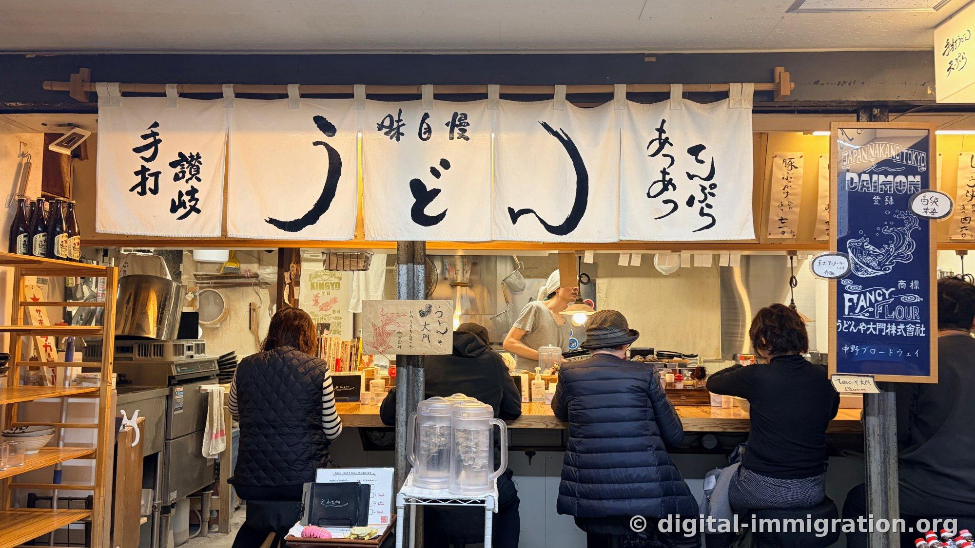 『手打 讃岐 うどん 味自慢 てんぷらあつあつ』と書かれた白いのれんの下で、カウンターに背を向けて座る客4名の店全景