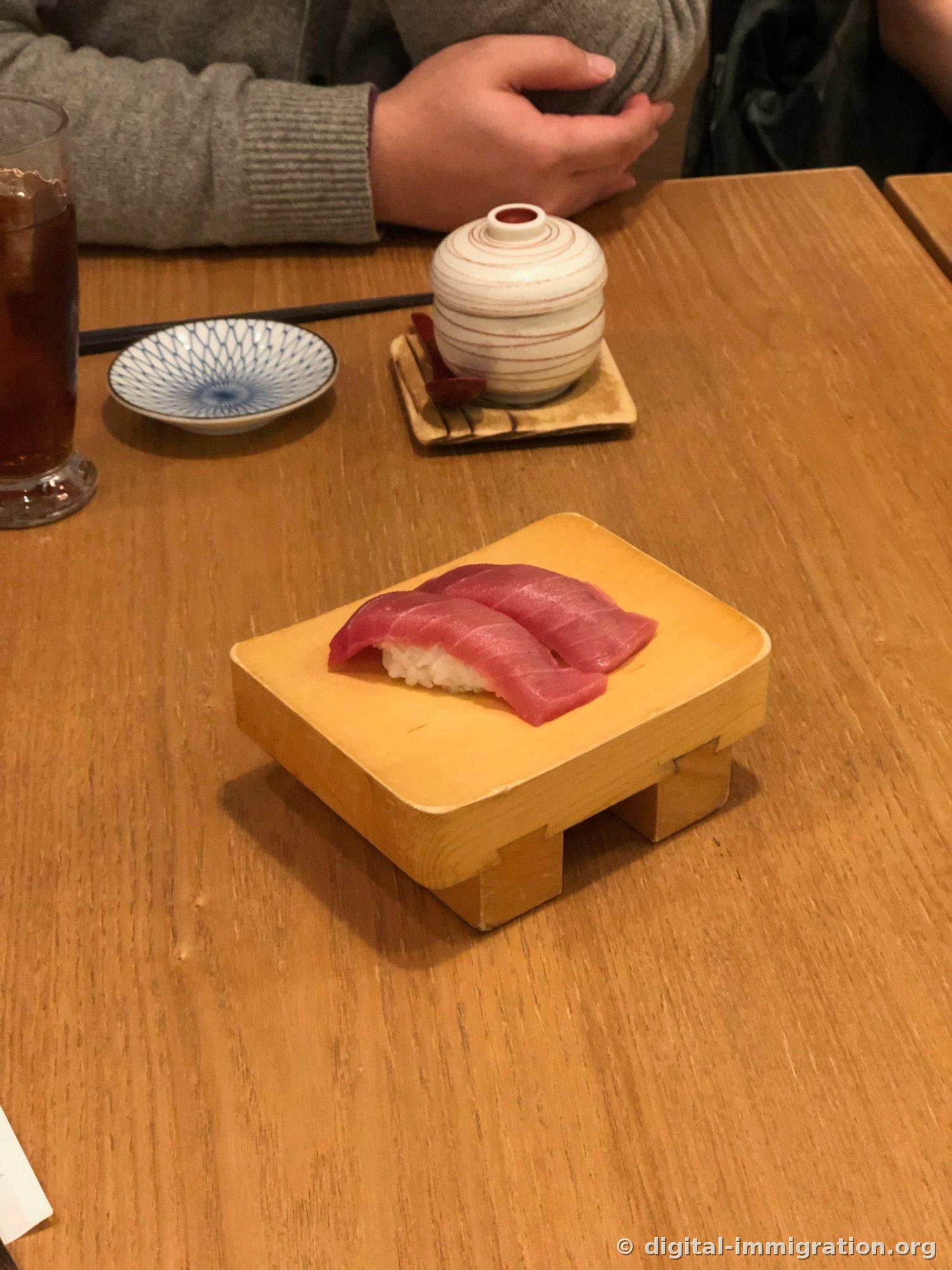 Deux pièces de nigiri de thon sur un support en bois, avec un bol couvert d'omelette à la vapeur derrière, un bol de sauce soja, et un verre de thé glacé