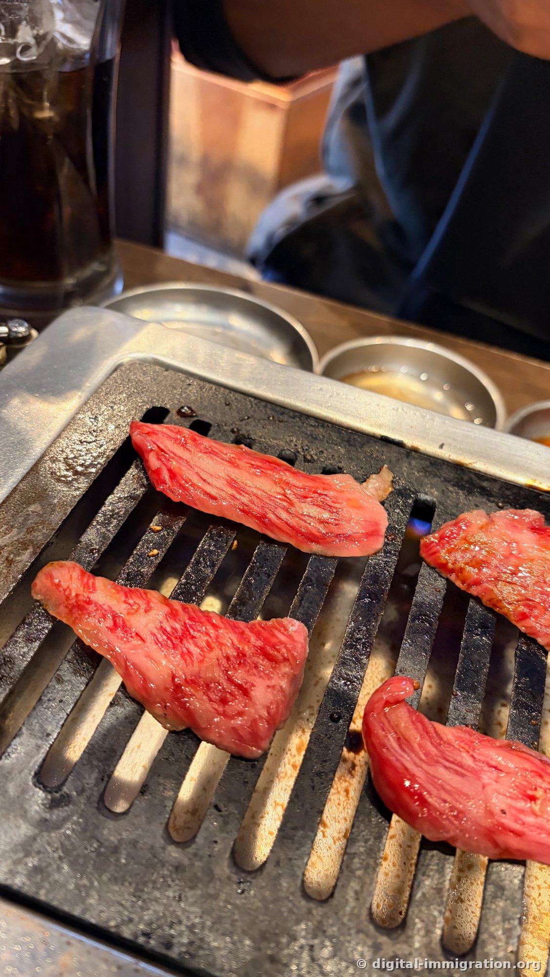 Four marbled slices of red beef on the wire mesh grill of a stainless-steel roaster; sauce dishes visible in the background