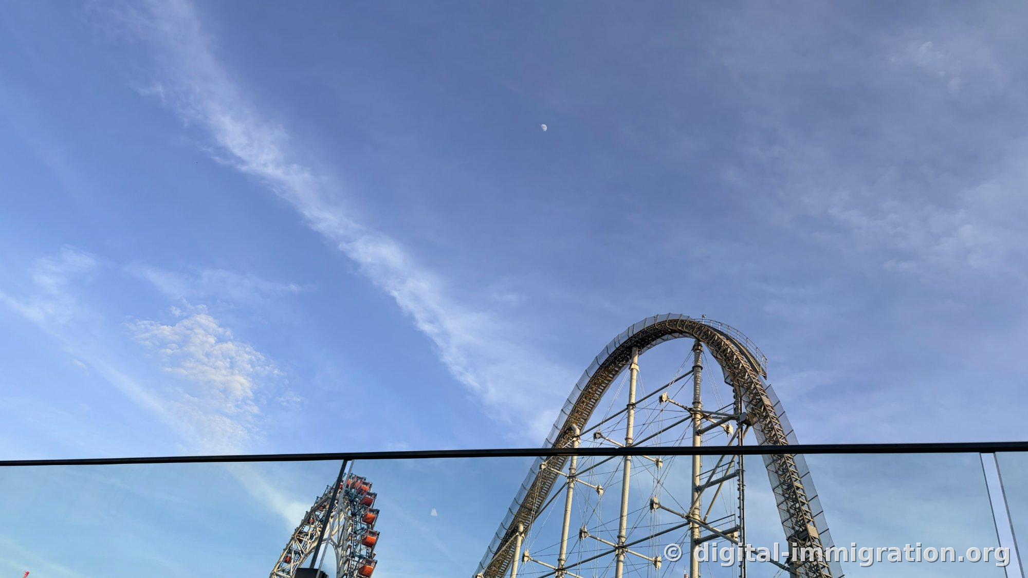 屋上から見上げた青い空、昼間の月と薄い雲、右にローラーコースター軌道の頂点と左下に観覧車のゴンドラ