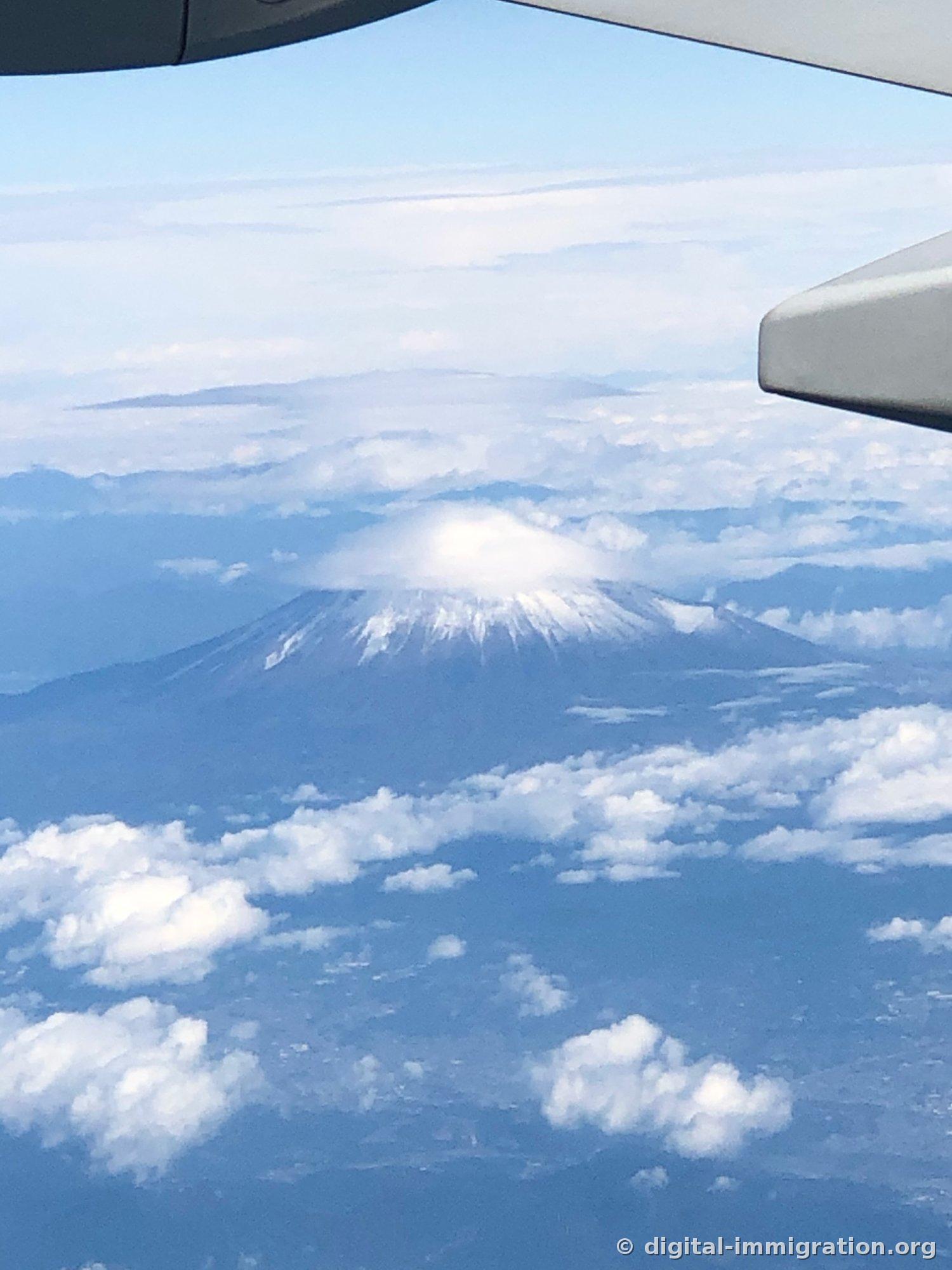 Une vue par la fenêtre avec le bord de l'aile de l'avion dans le coin supérieur droit, montrant le Mont-Fuji coiffé d'un épais nuage en forme de chapeau au sommet et des nuages blancs épars en dessous
