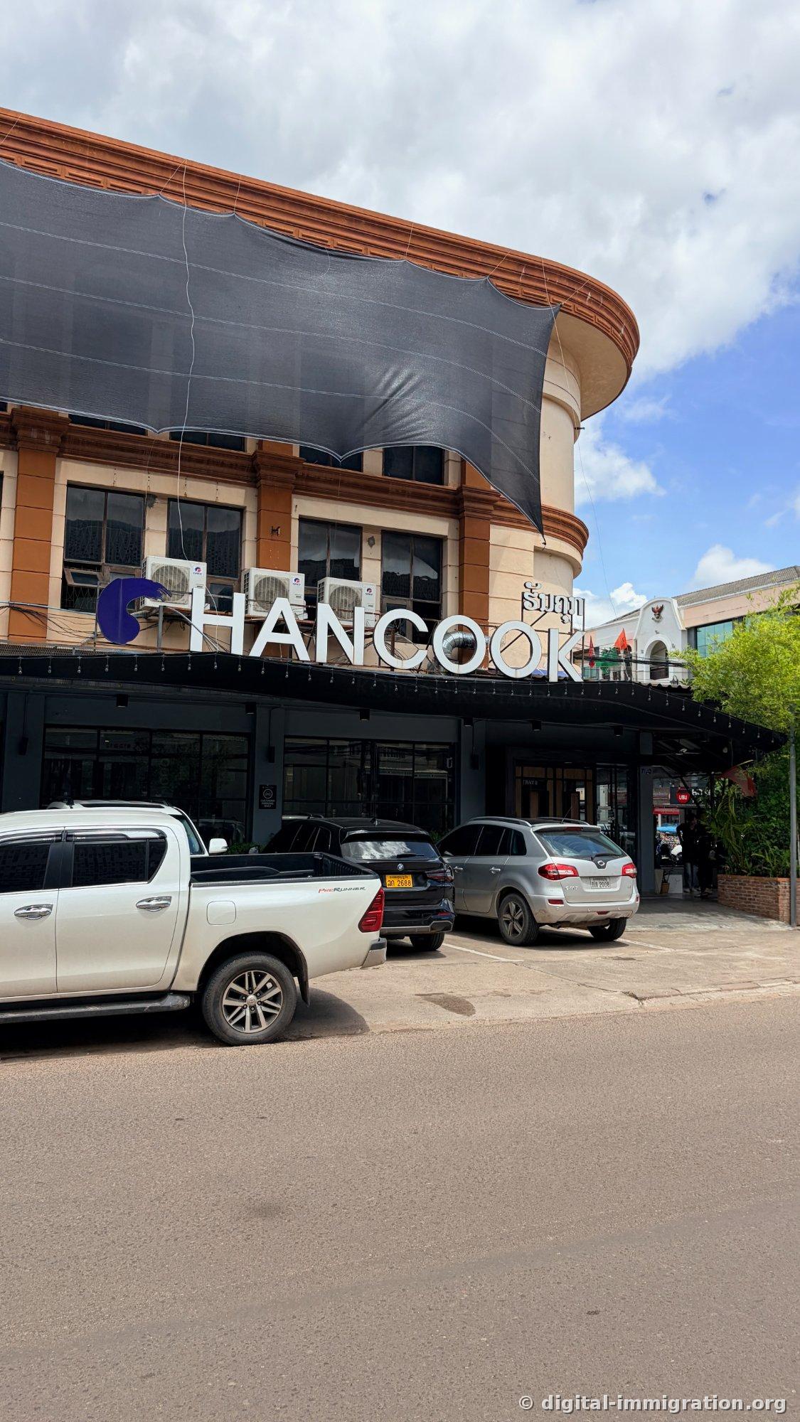 Rez-de-chaussée d'un bâtiment à étage aux moulures crème et orange, avec l'enseigne HANCOOK en grandes majuscules blanches et une enseigne en laotien à côté ; le premier étage est recouvert d'un store noir occultant, et un pick-up blanc ainsi que d'autres voitures sont garés devant.
