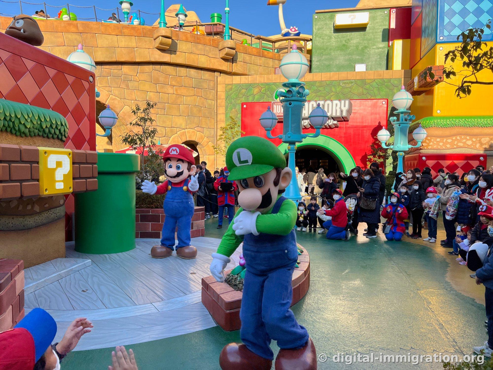 Mario and Luigi wave to the audience on a circular stage in front of the Toad Factory sign, surrounded by masked guests