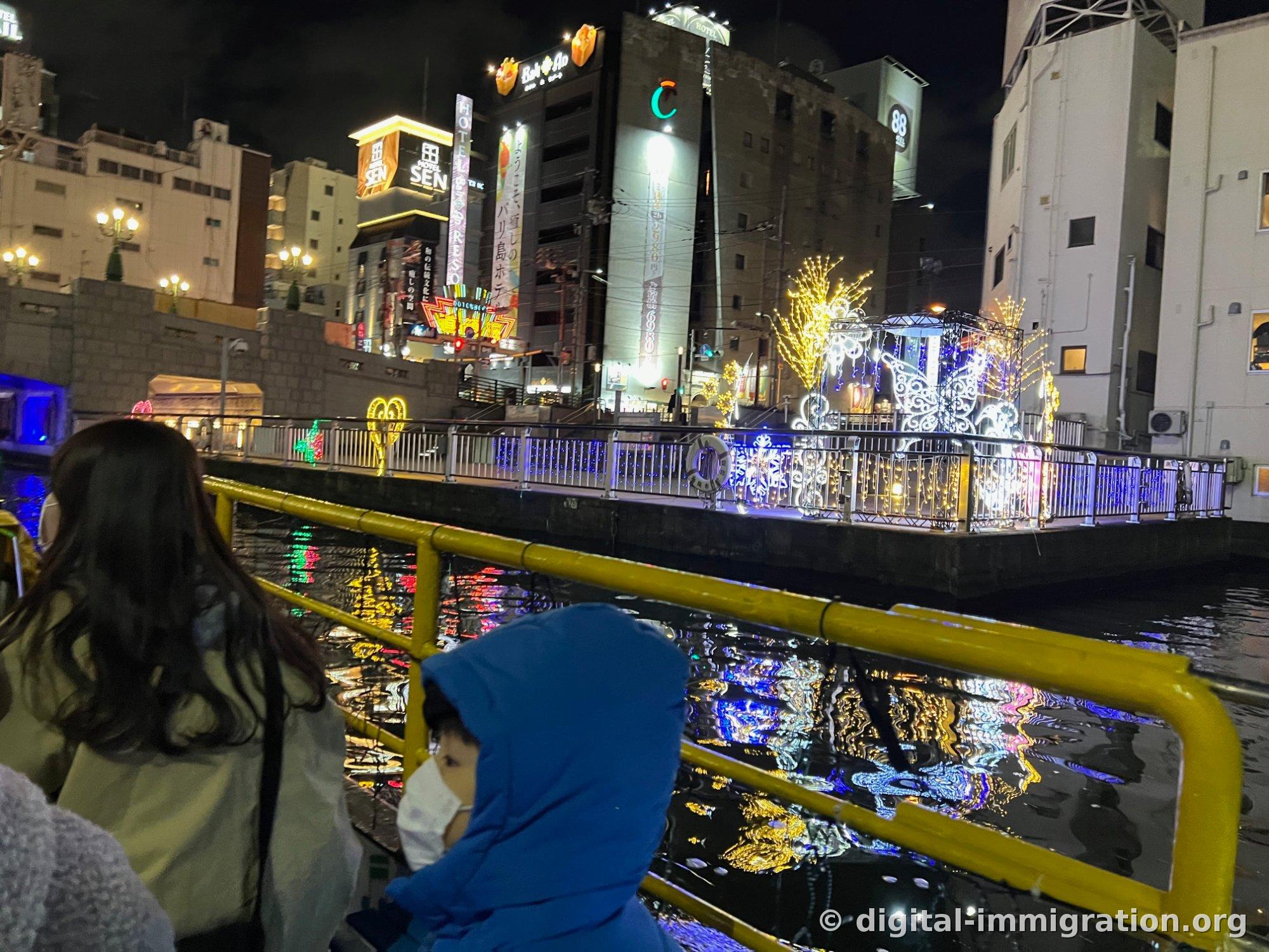 Installation d'illumination en forme de lampes blanches et jaunes sur la promenade le long du fleuve, enseigne hôtel SEN derrière, reflet de la lumière dans l'eau
