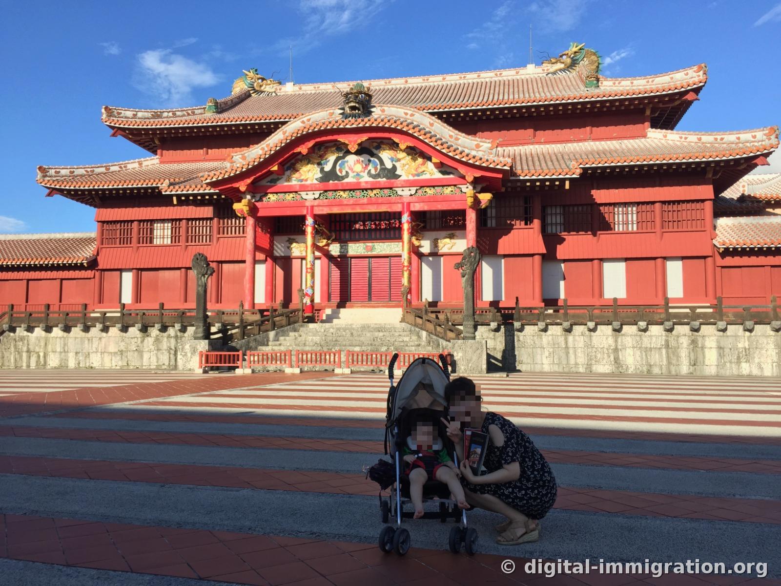 Full view of Shuri Castle's vermillion Seiden beyond Unaa plaza with its red and white striped floor, a child in a stroller in the foreground