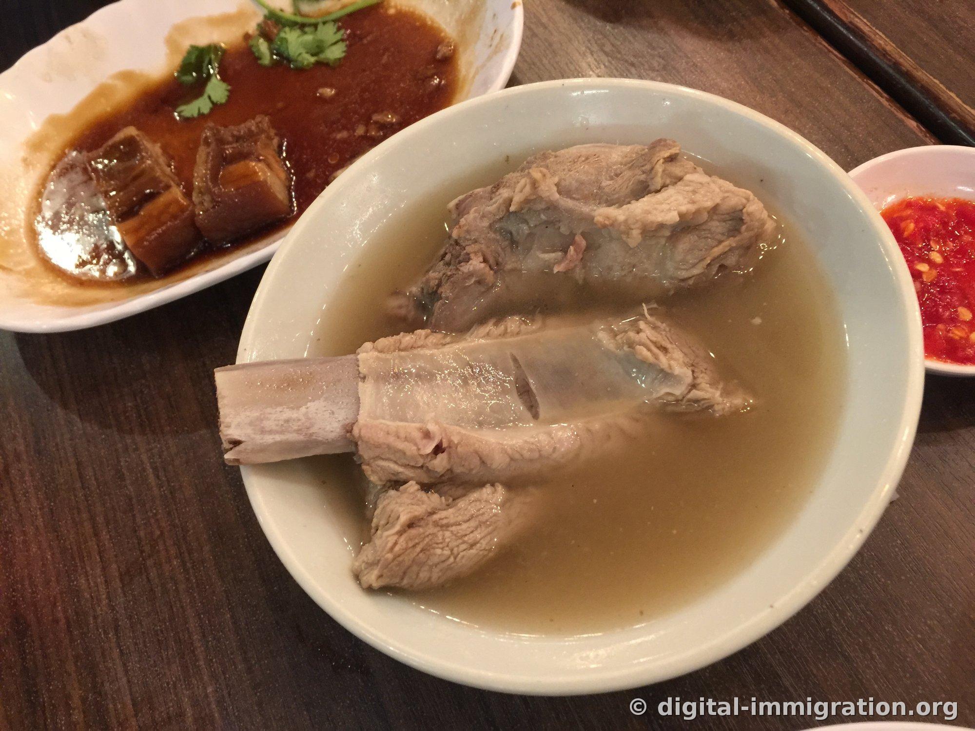 Un bol de Bak Kut Teh avec un bouillon clair épicé, accompagné d'un plat de viande braisée à la sauce soja et d'une sauce de piment haché rouge