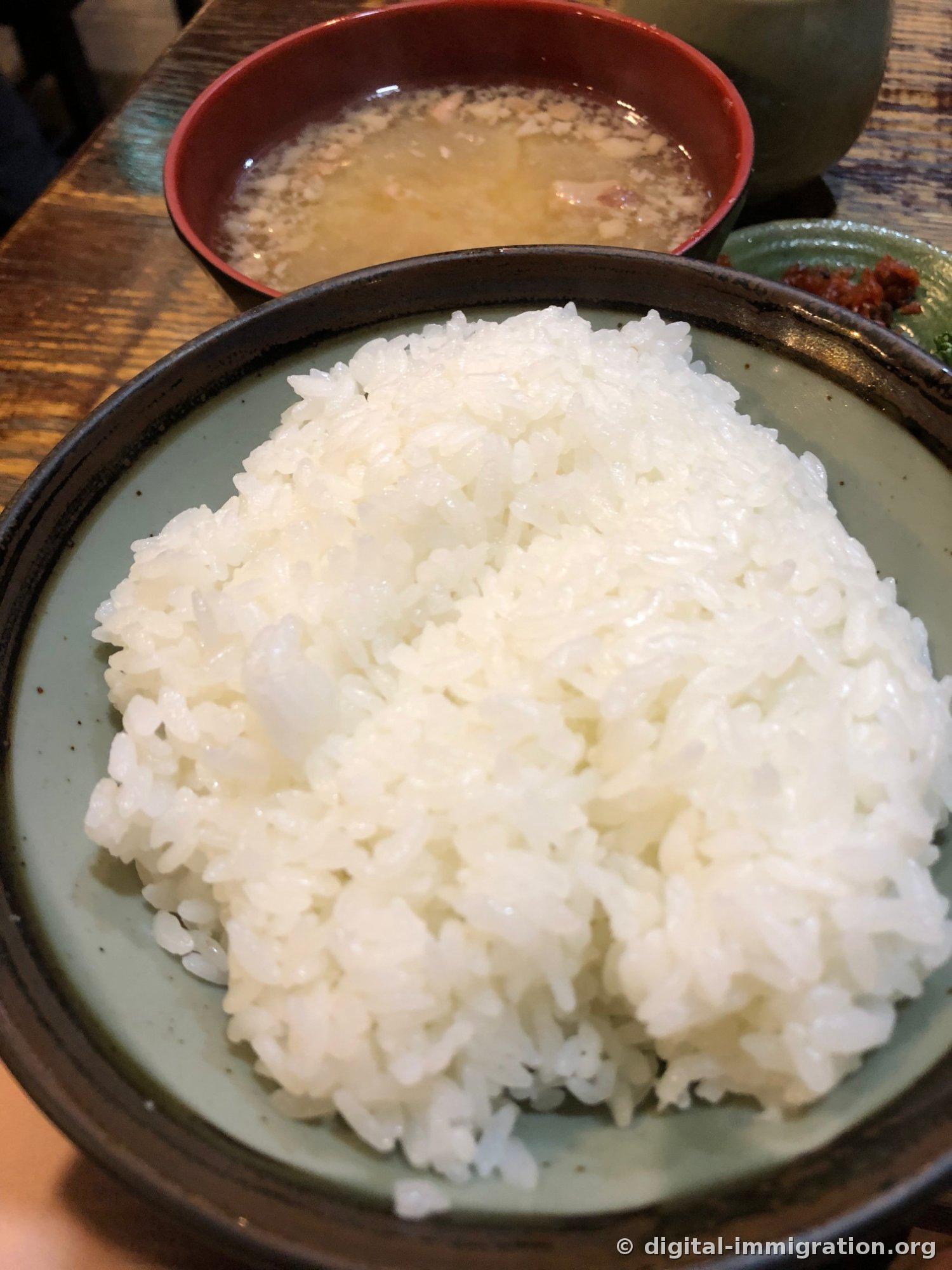 A large bowl of heaping white rice and, behind it, a red lacquered bowl containing pork miso soup, with red pickles on the right