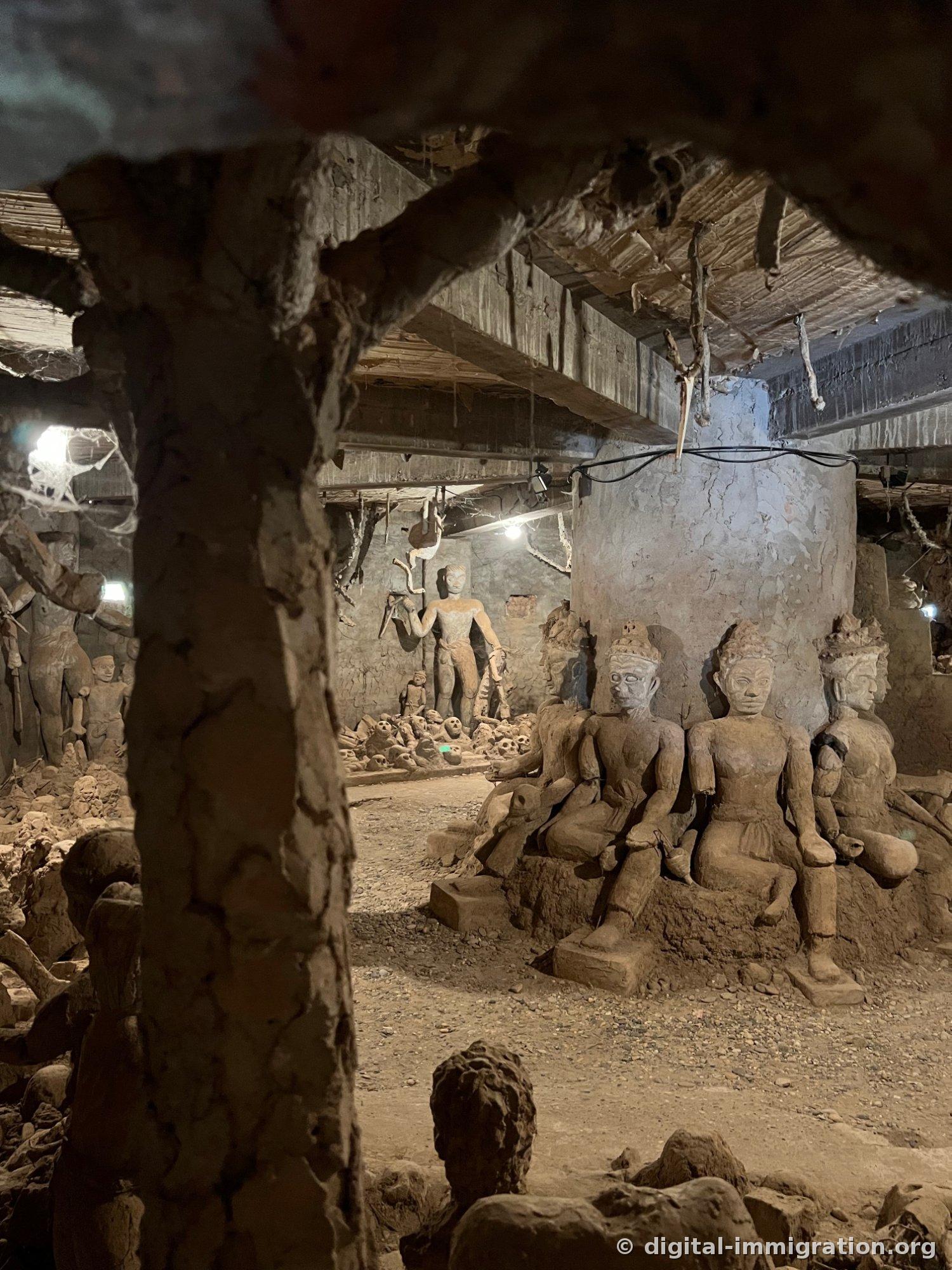 Trois statues de Bouddha assis visibles derrière un pilier, avec des sculptures de crânes au sol et des formations de racines suspendues du plafond