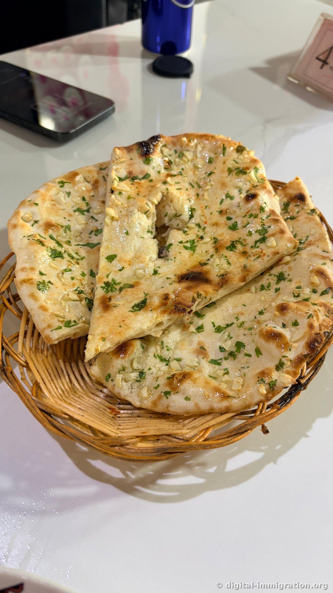 Trois morceaux de naan à l'ail empilés dans un panier en rotin. L'ail haché et la coriandre sont saupoudrés dessus, et les bords montrent des marques de brûlure du four tandoori
