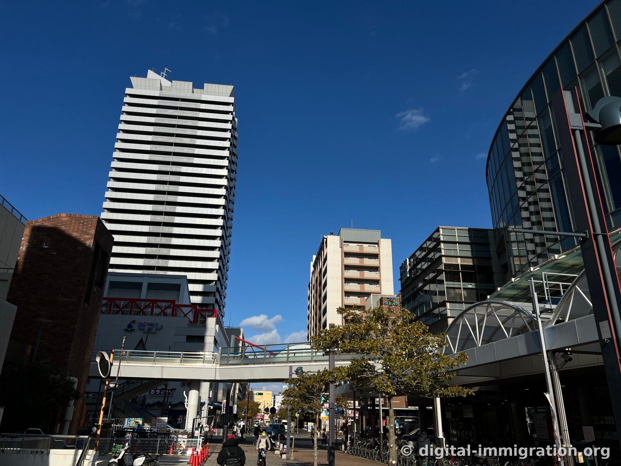 Sous un ciel bleu, des immeubles d'habitation de grande hauteur, l'enseigne Pifre et une passerelle piétonne qui traverse la rue devant la gare Shin-Nagata