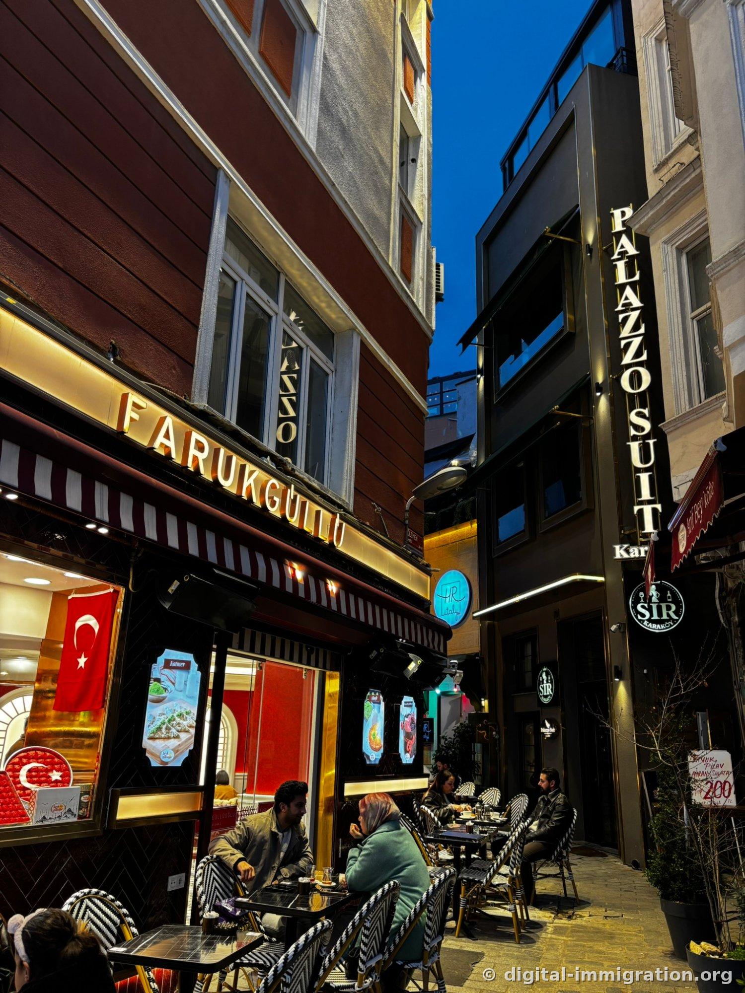 Outdoor café tables in Karaköy alley at dusk with the golden FARUKGÜLLÜ sign and vertical PALAZZO SUIT sign visible under blue sky