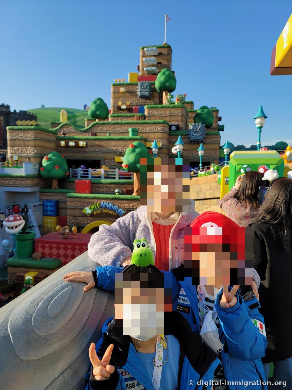 Children and an adult wearing Yoshi plush hats and Mario hats pose against a Yoshi Adventure sign and tiered hills background, leaning on the railing with peace signs