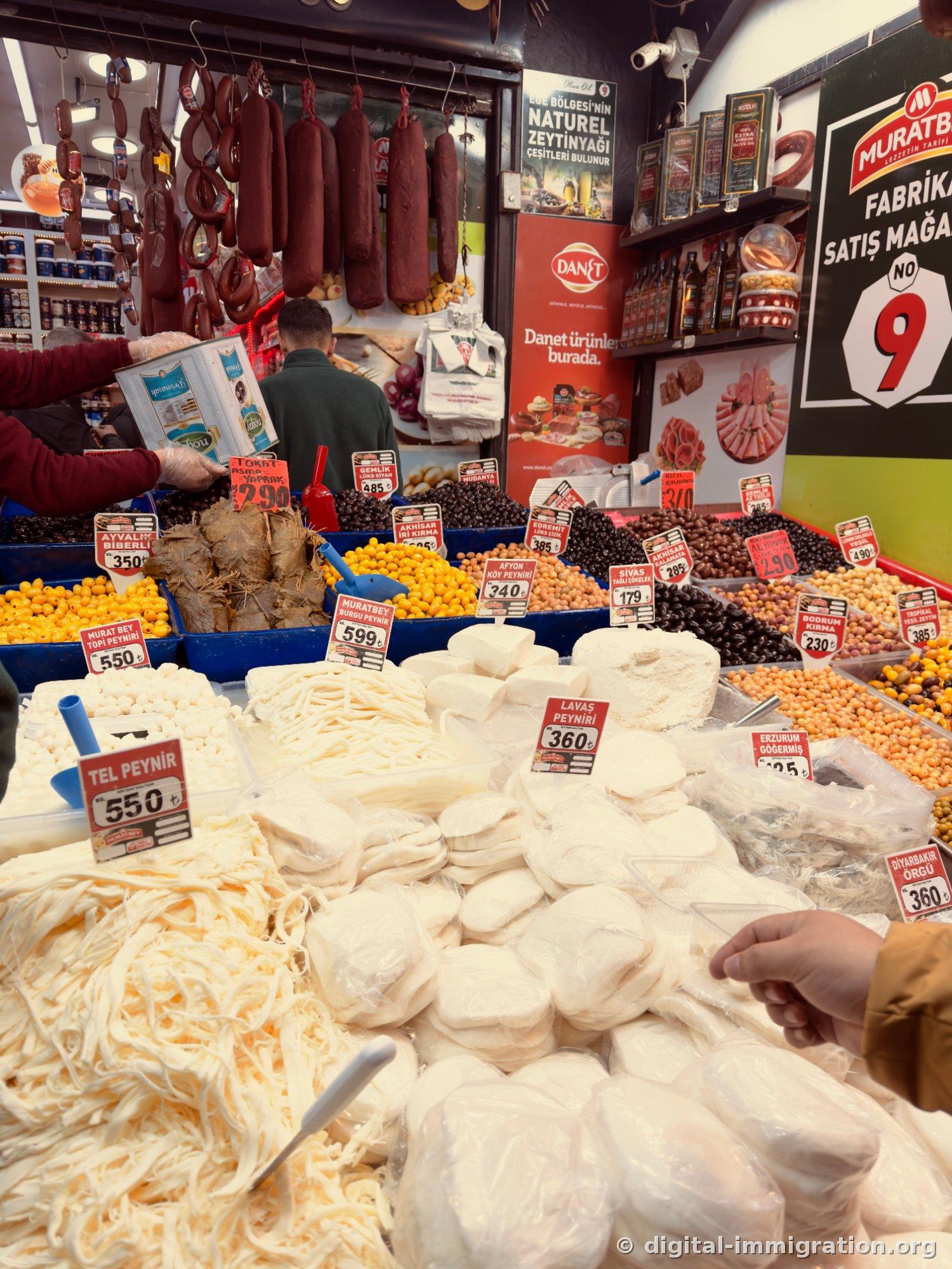 Soudjouk sausages hanging from the ceiling, piles of white cheese and various olives in boxes below, red price signs like TEL PEYNİR 550, LAVAŞ PEYNİRİ 360 stuck in