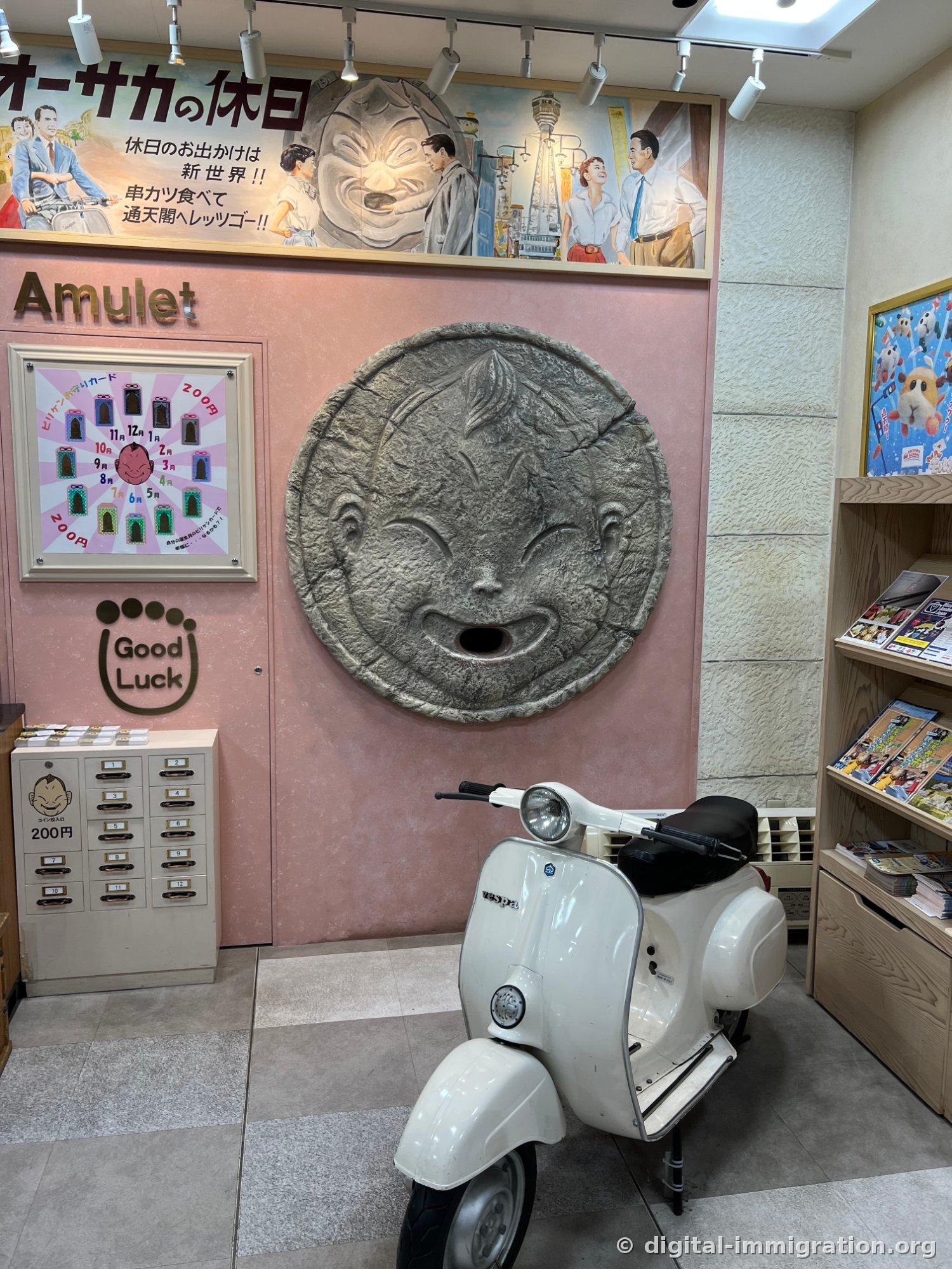 A large Billiken face relief stuck to a pink wall, with a white Vespa scooter standing in front