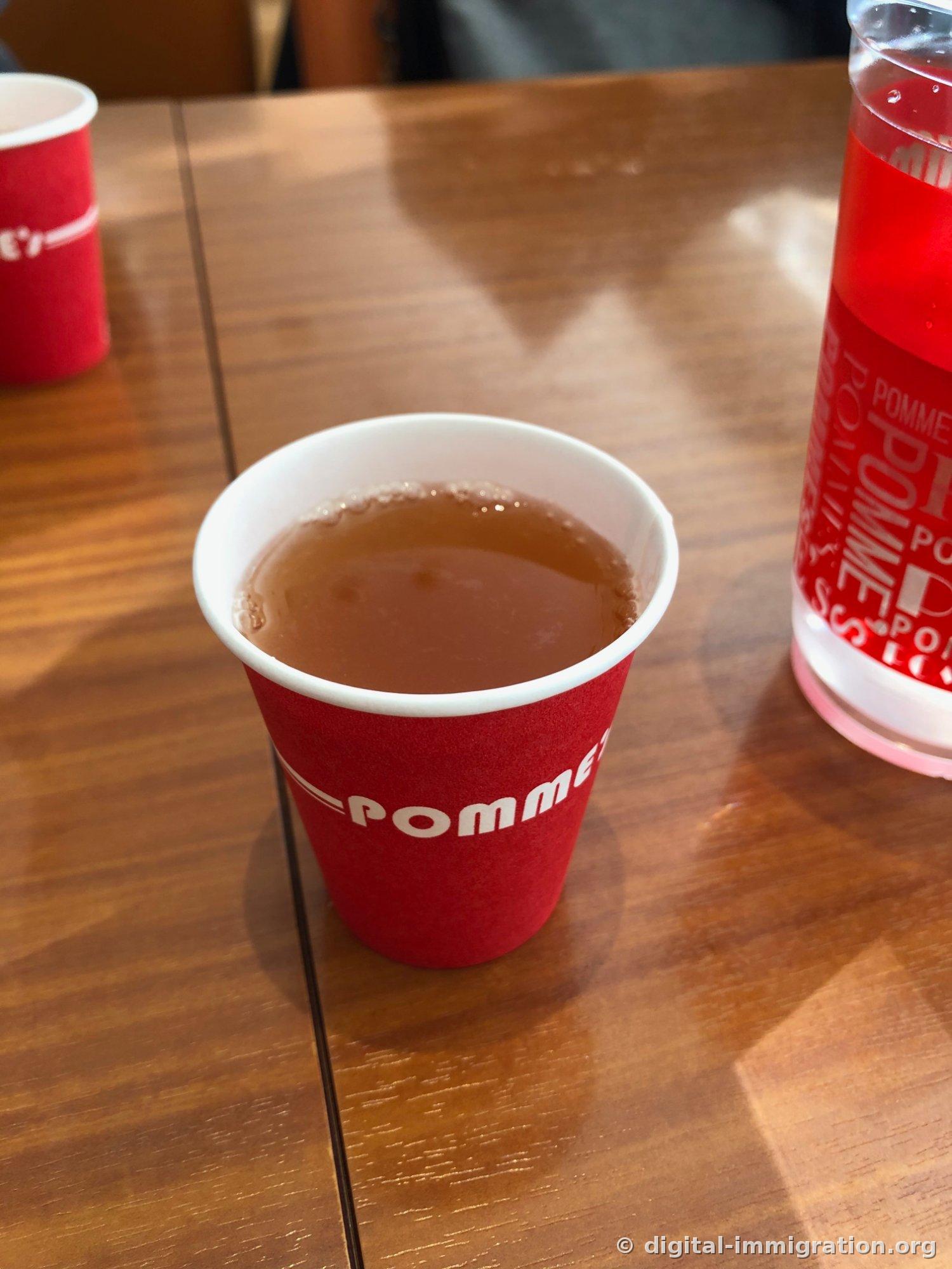 Soupe miso servie dans un gobelet en papier avec le logo rouge POMME's sur une table en bois, à côté un verre avec une boisson rouge