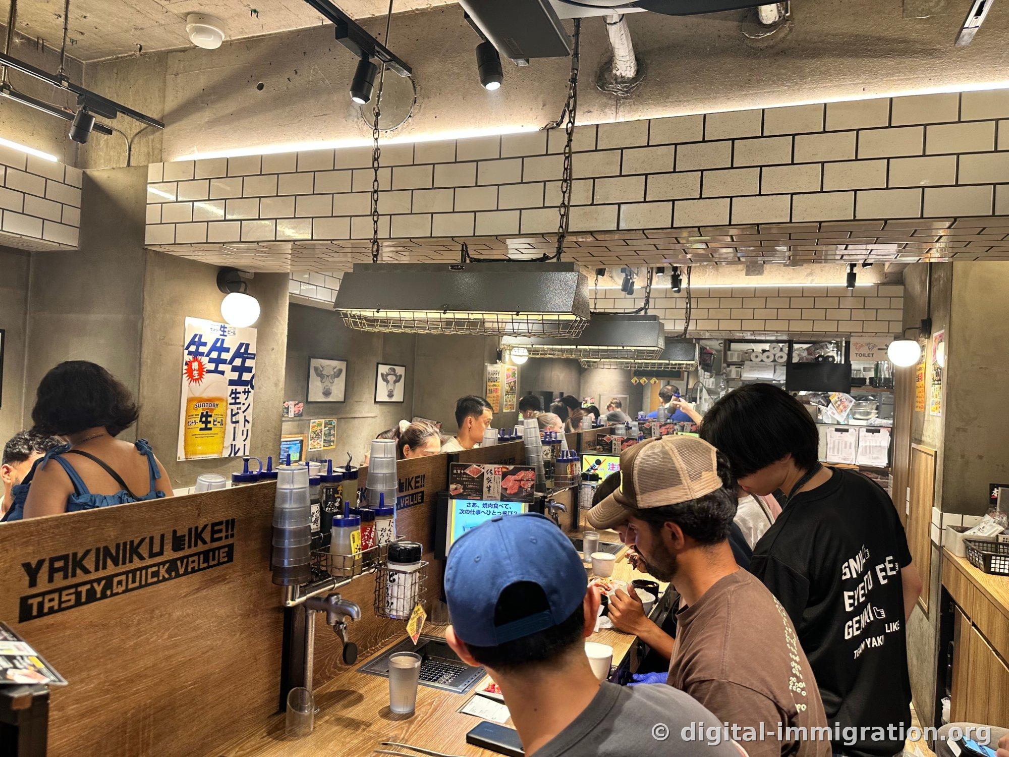 L'intérieur du restaurant Yakiniku Like à Shibuya. Murs carrelés blancs et plafond en béton apparent, avec une hotte d'extraction suspendue par des chaînes au-dessus du comptoir où les clients font griller leur viande chacun de leur côté