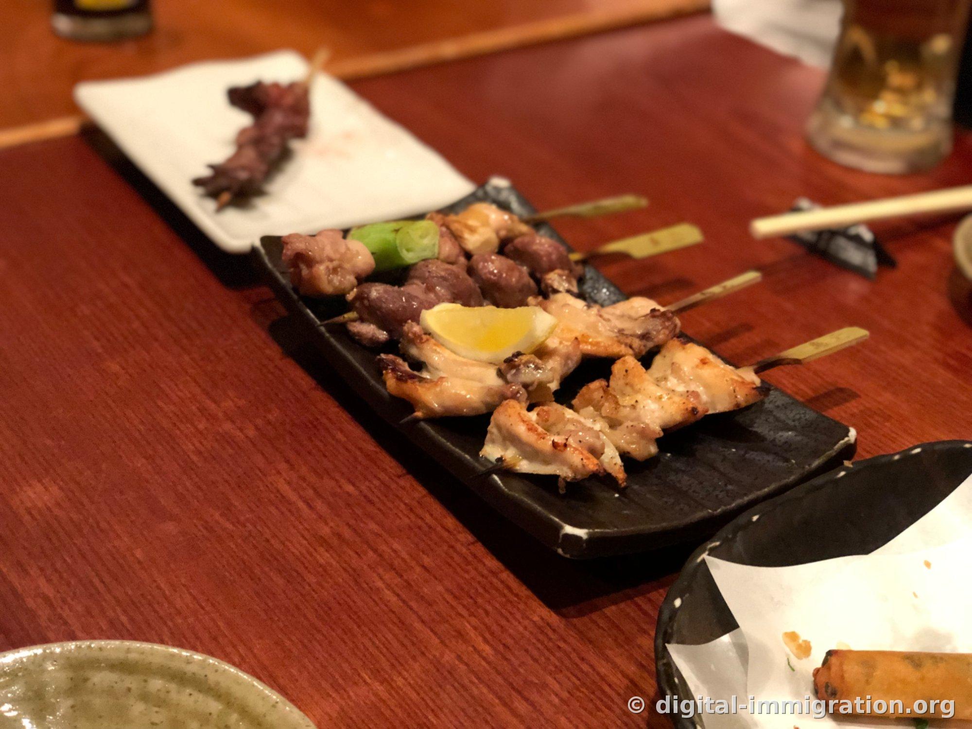 黑色长方形盘子里盛着葱间串、鸡心和鸡肉串，以及一片柠檬，旁边的白盘子里有吃完后的竹签