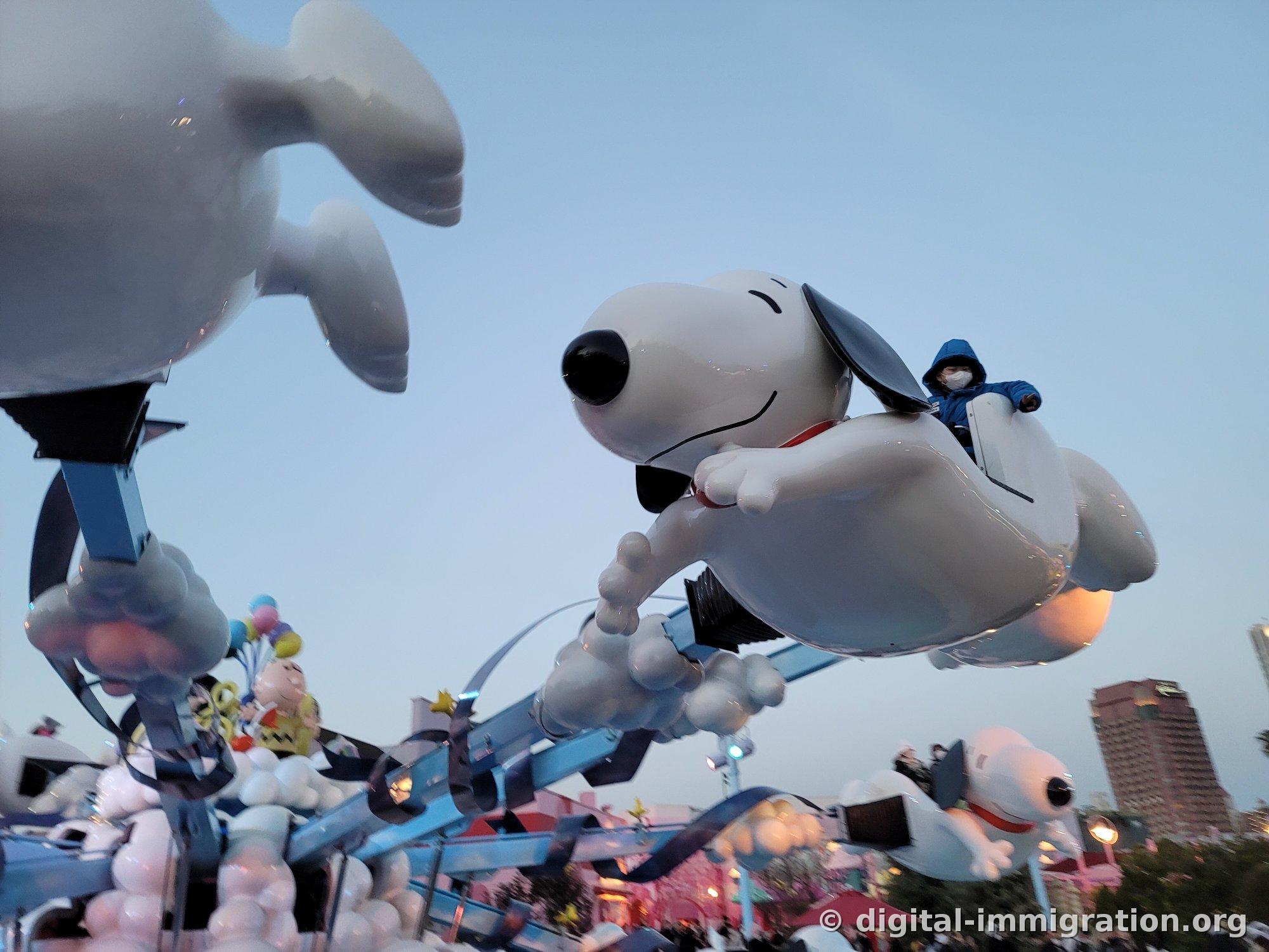 Large Snoopy character ride flying across the sky at dusk with a child in blue jacket on top, Osaka city buildings visible in the background