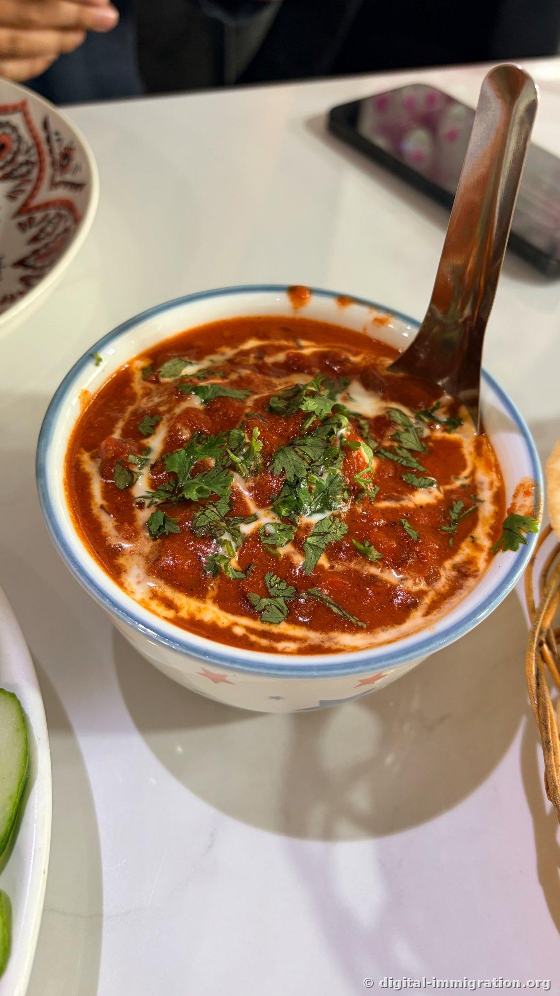 Curry à base de tomate rouge dans un bol en céramique à bordure bleue. La crème fraîche forme un tourbillon à la surface, garni de coriandre, avec une louche en acier inoxydable plantée dedans