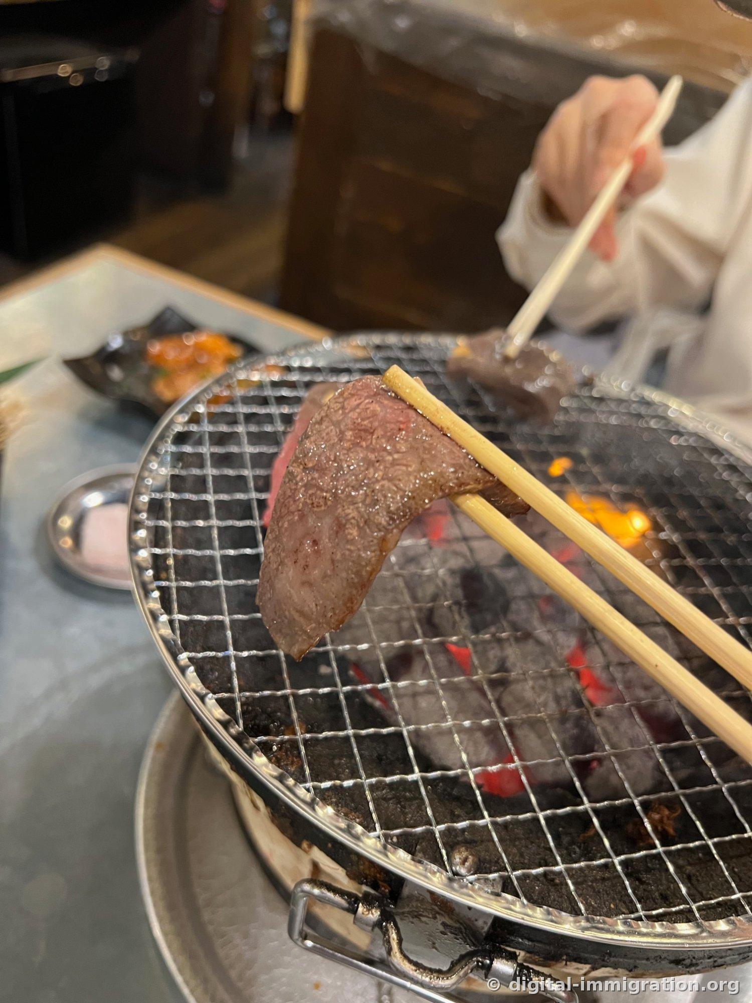 木箸で網の上の焼かれた肉一点を持ち上げている様子。後ろに黒いお皿に入ったおかずと金属のお皿が見える