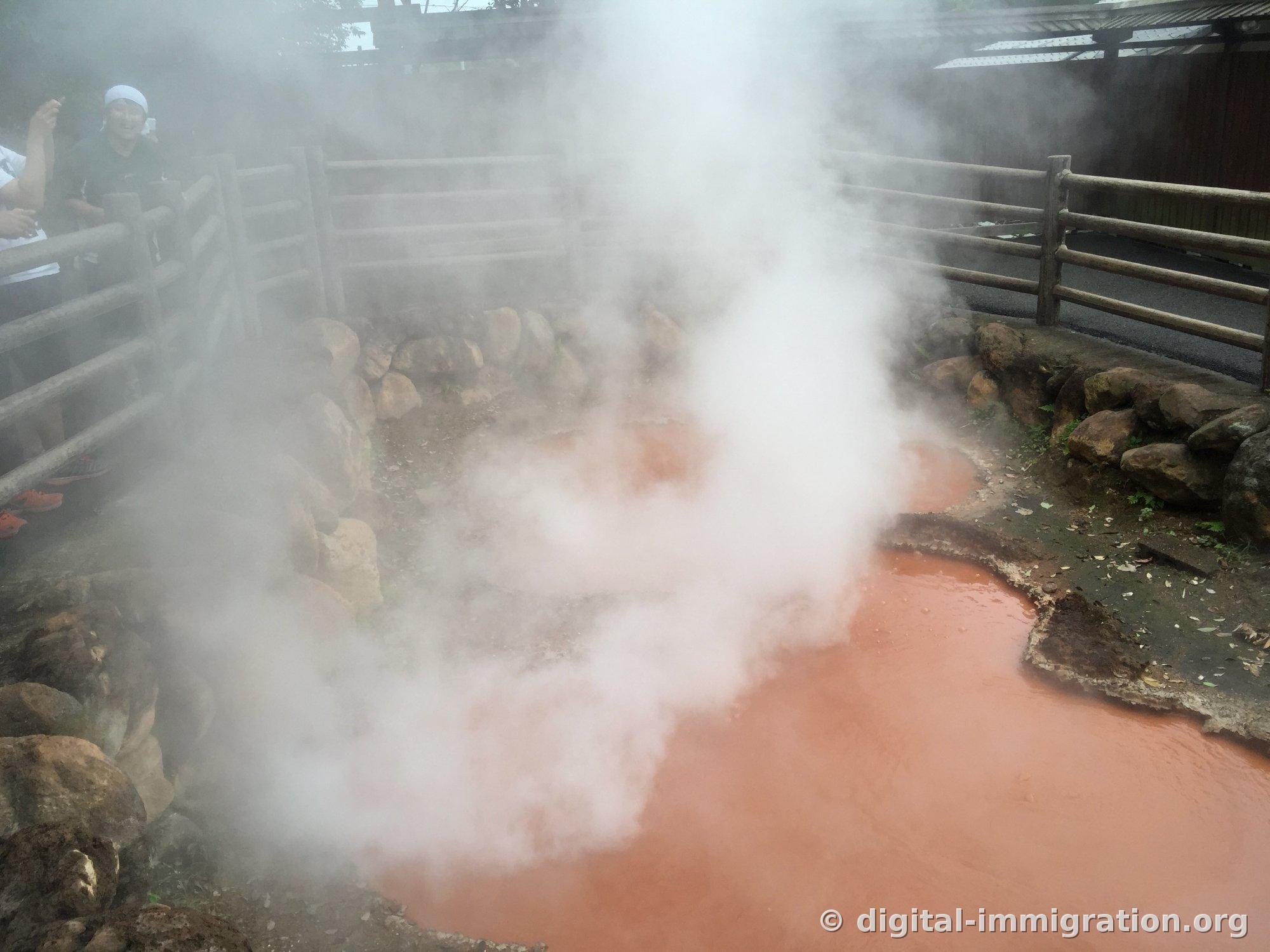 Un bassin d'eau chaude de couleur brique rougeâtre avec une colonne de vapeur blanche s'élevant verticalement, des spectateurs visibles au-delà de la barrière en bois