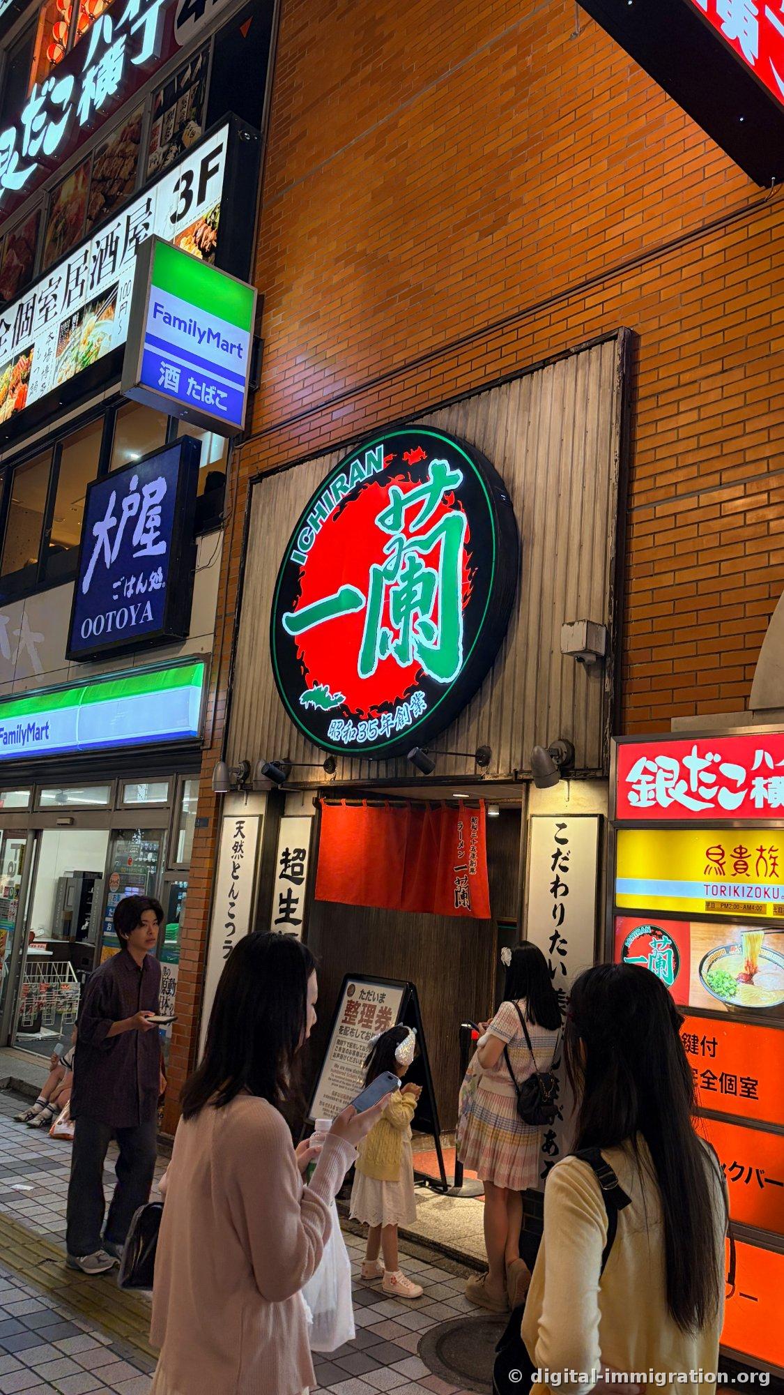 La nuit, devant le panneau de distribution des numéros de ticket, des gens attendent dans la rue éclairée par le néon. On voit aussi les enseignes de Kindako Yokocho et Torikizoku de l'immeuble à côté.