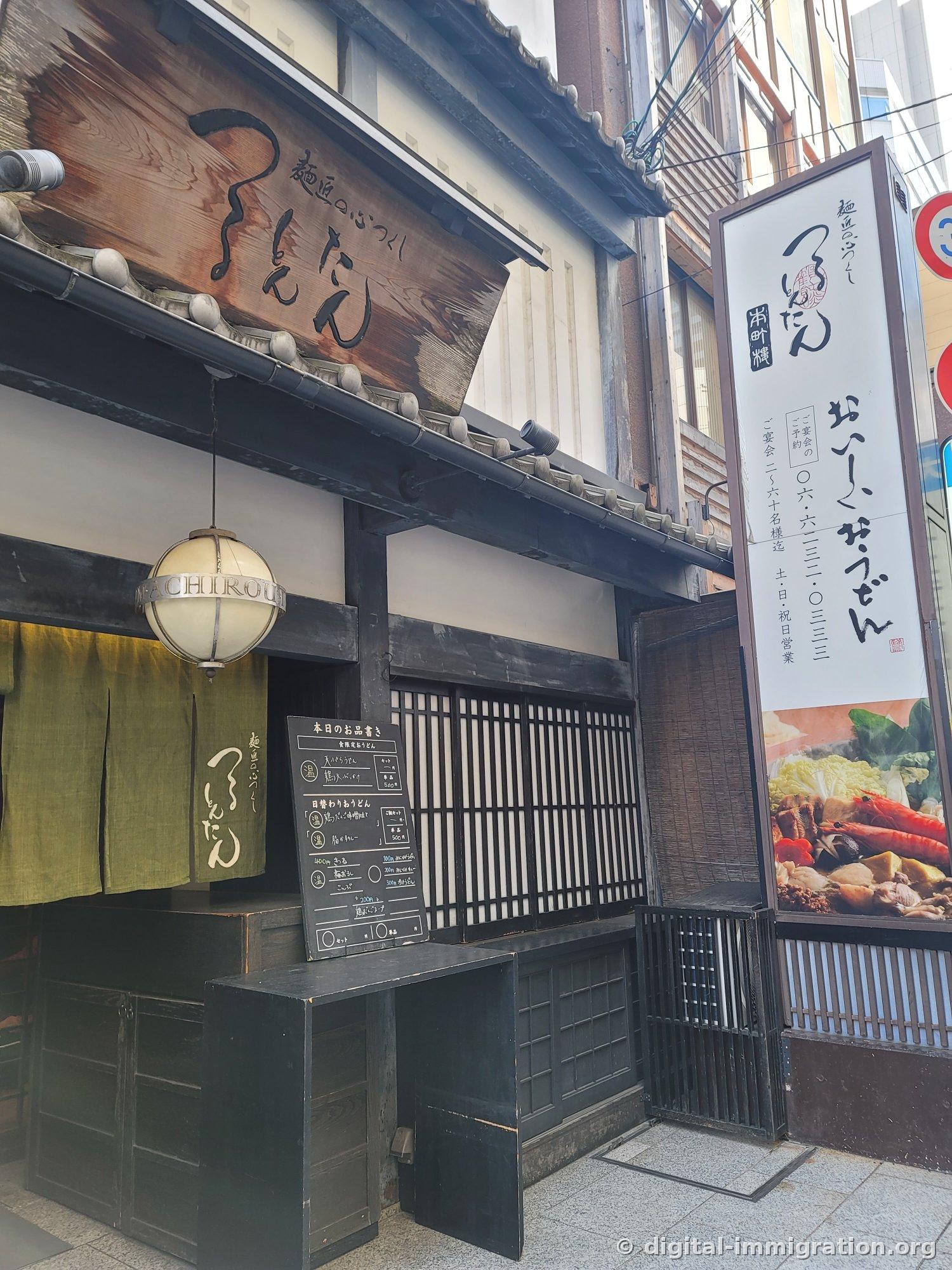 L'entrée de la succursale Honmachi avec le panneau du restaurant Tsurutontan écrit au pinceau sur bois sous l'avant-toit aux tuiles, le rideau vert clair (noren), et une ardoise avec le menu manuscrit devant l'entrée
