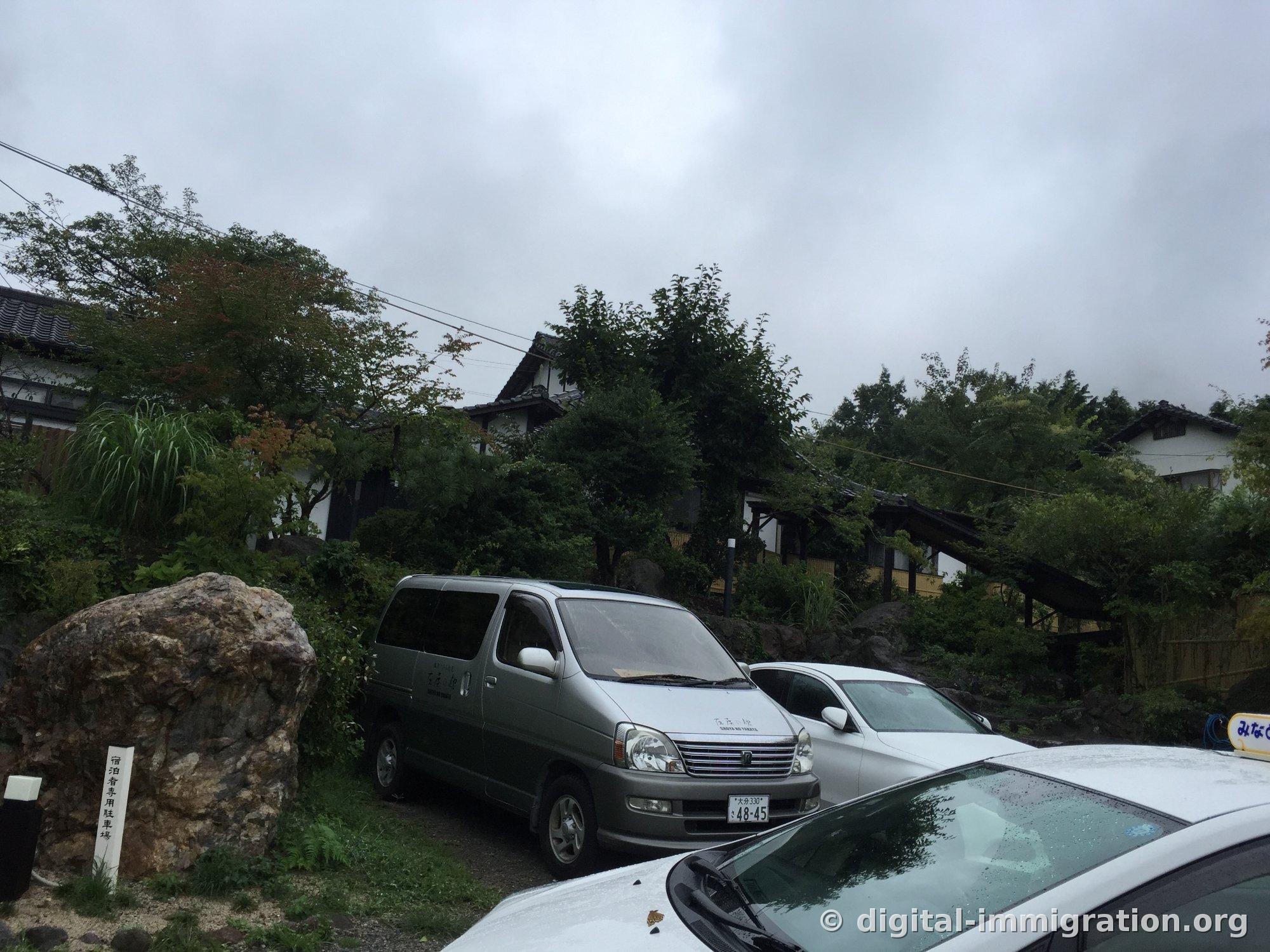 Le monolithe du parking réservé aux clients et une camionnette grise avec le logo de 庄屋の館, avec une maison de tuiles sur la colline