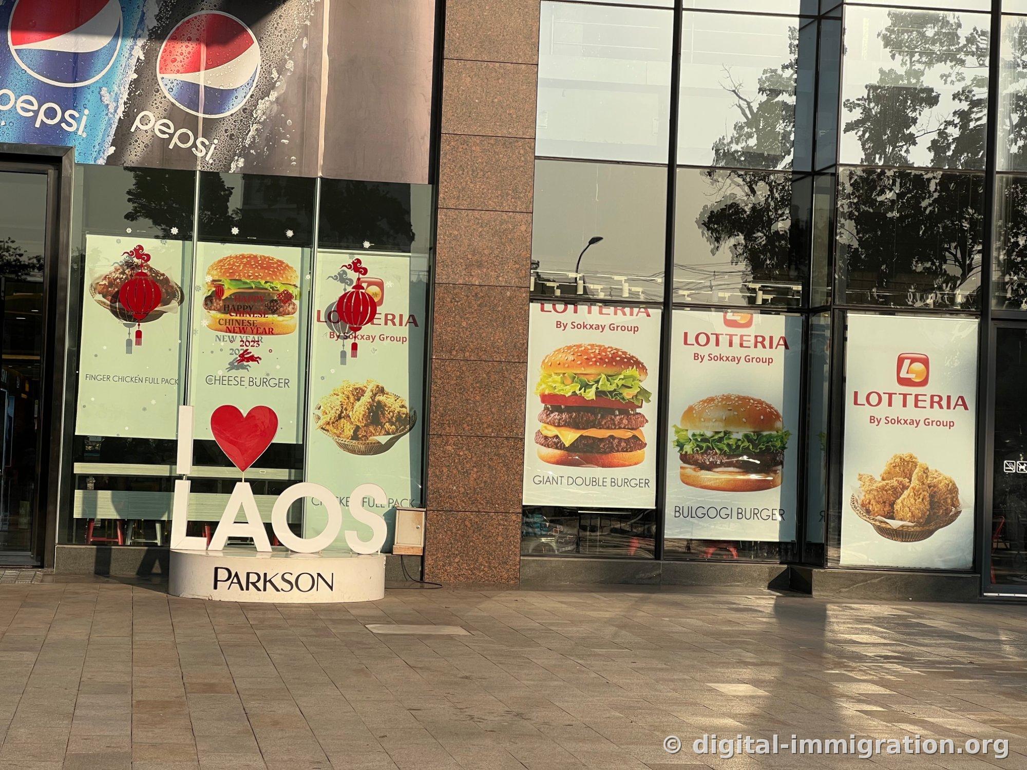 Publicités de burgers Lotteria sur la façade du premier étage du Parkson Mall avec la structure blanche I LOVE LAOS PARKSON, et une publicité Pepsi au-dessus