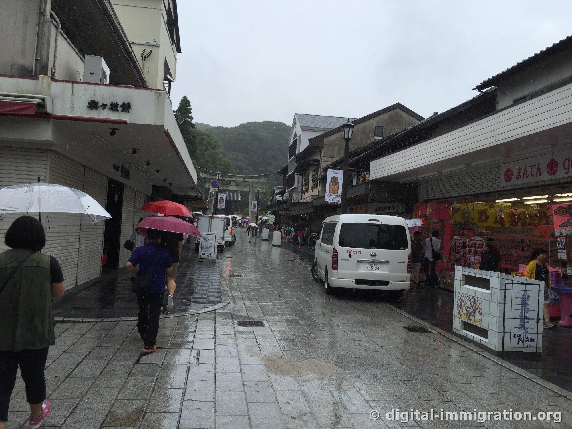 Dazaifu Sando in the rain, wet stone pavement and people with umbrellas, shop sign reading '梅ヶ枝餅' visible on the building to the left