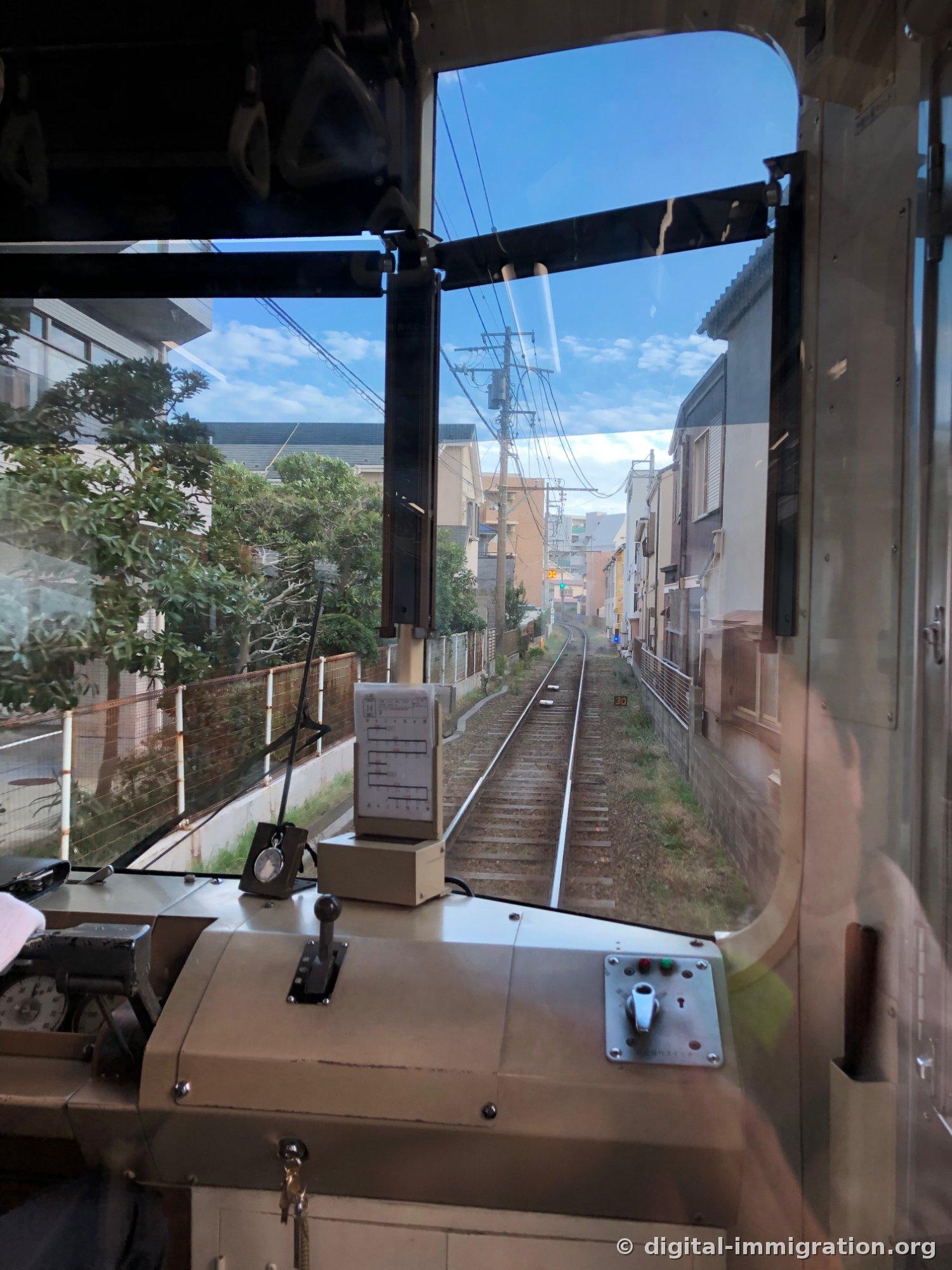 Au-delà de la vitre du premier wagon, une voie unique traversant un quartier étroit de maisons avec un panneau de limitation de vitesse 30 visible sur la droite